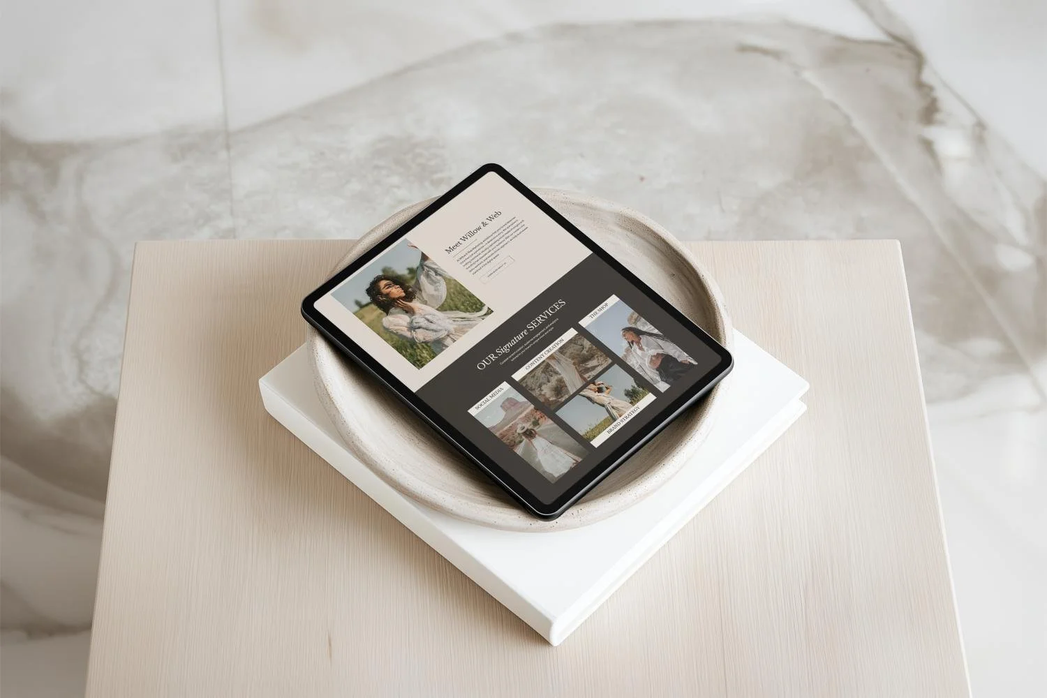 A tablet displaying a website with images and text, placed on a white ceramic bowl on top of a white book on a light-colored table.