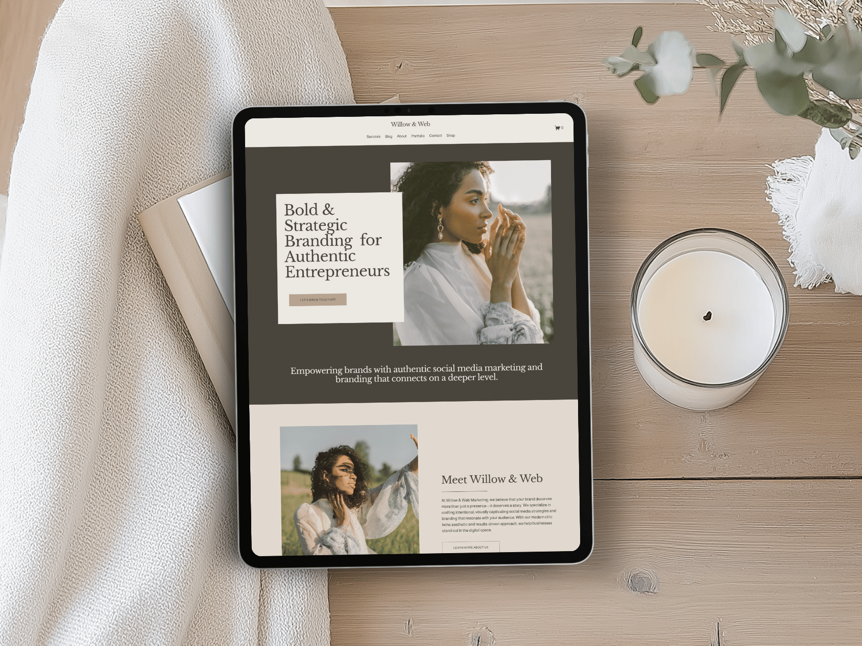 An iPad displaying a website about authentic branding for entrepreneurs, placed on a wooden table next to a lit candle and a white cloth with a bouquet of dried flowers.