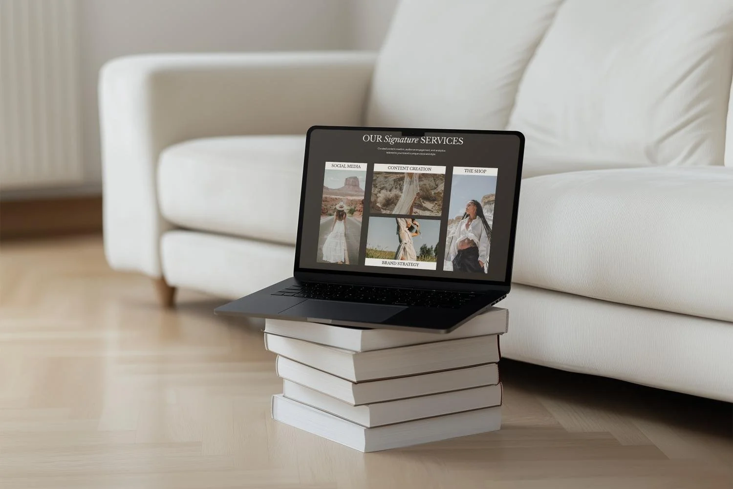 A laptop computer sitting on top of a stack of white hardcover books on a light wood floor, with a white sofa in the background.
