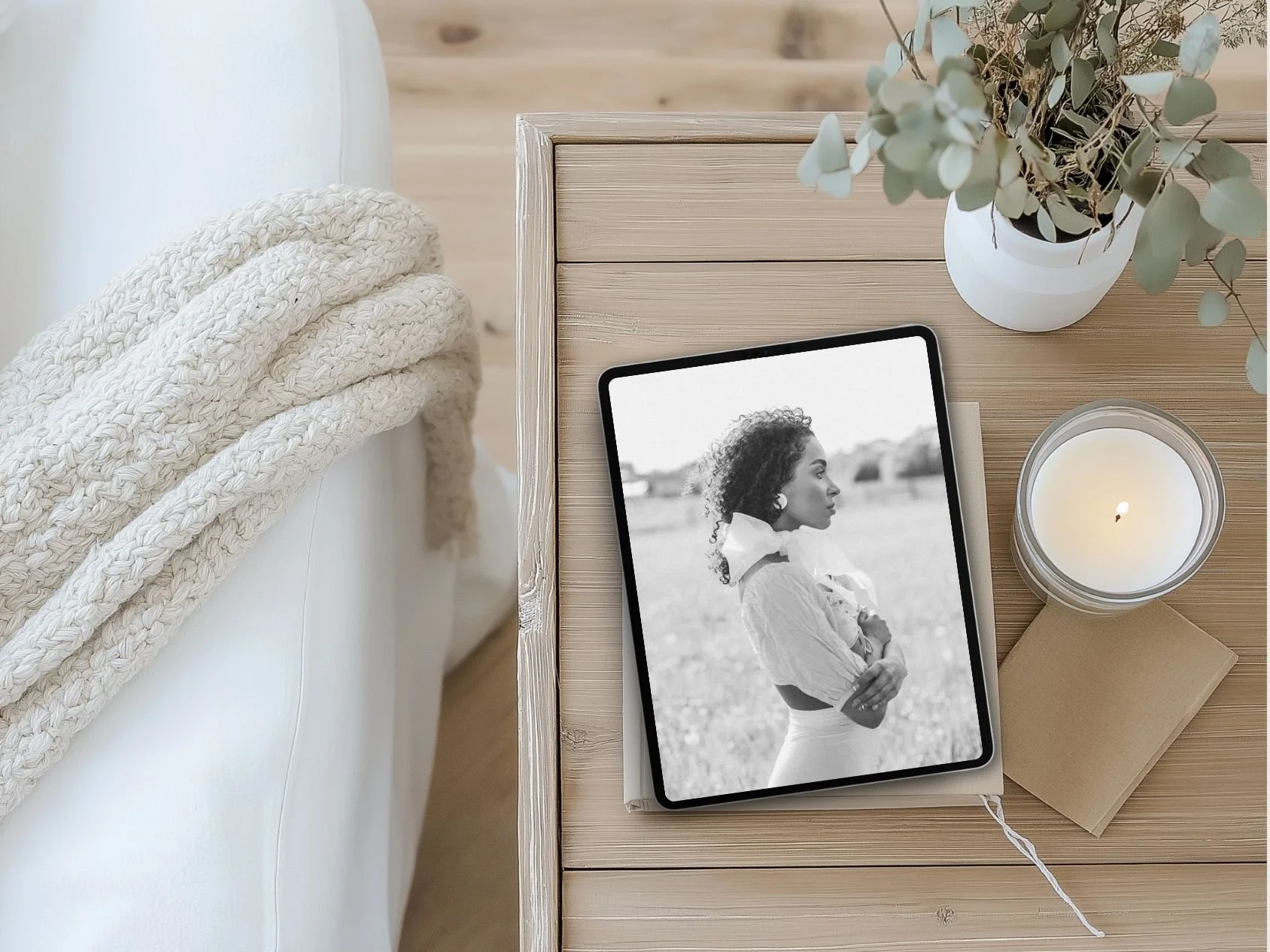 A wooden bedside table with a black and white photo of a woman, a lit candle in a glass jar, a white notebook, and a white potted plant with green leaves. A knitted white blanket is draped over the edge of a white bed nearby.