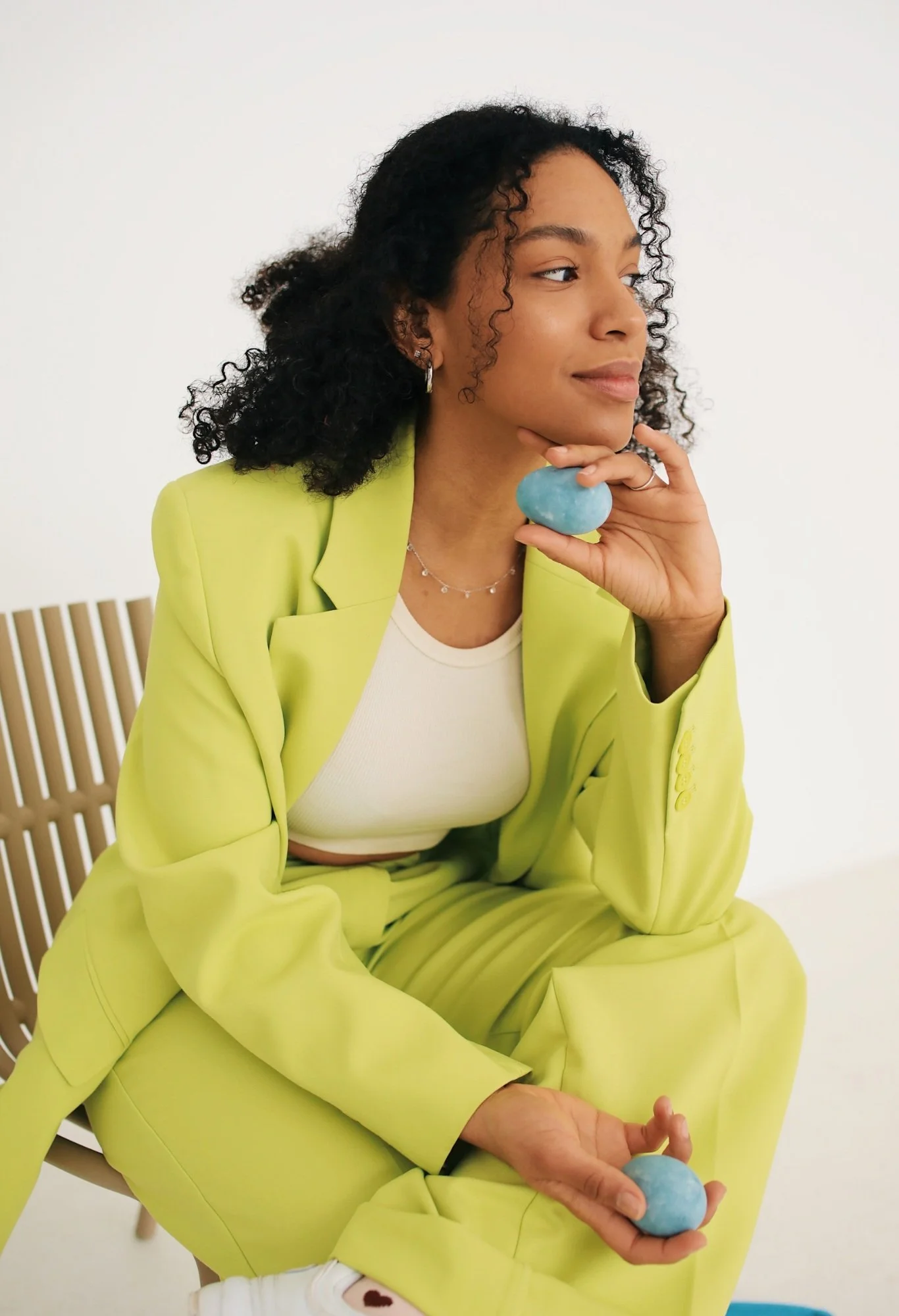 A woman with curly black hair, wearing a yellow suit and white top, sitting on a wooden chair holding a blue stone in her right hand near her face, with her left hand resting on her knee, in a minimalistic white room.