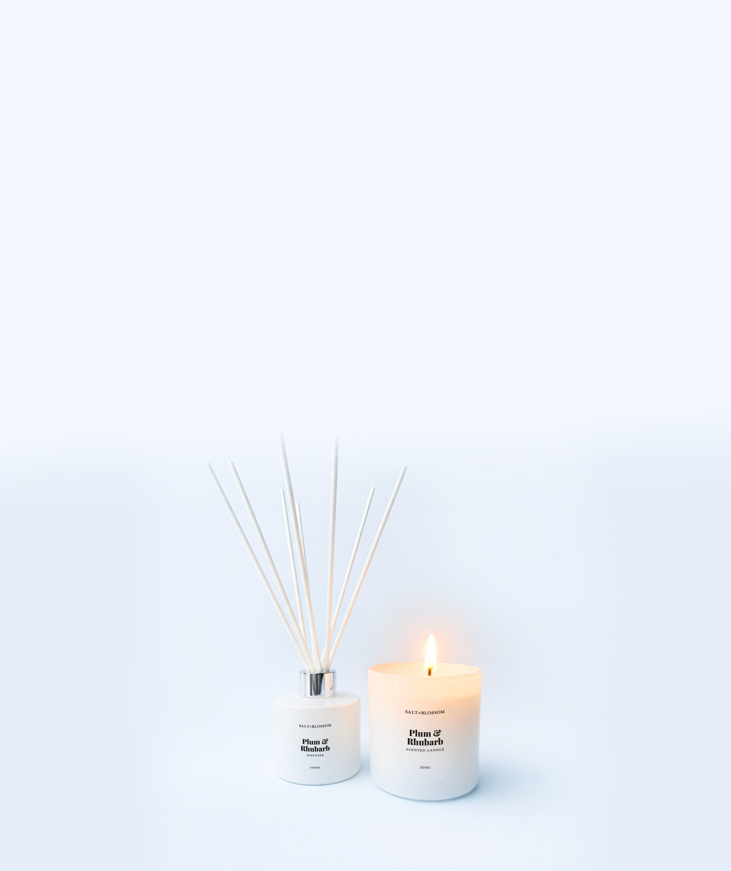 Three white candles, one lit, labeled 'Salt & Blossom' with scents 'Plum & Rhubarb' and 'Plum & Rhubarb', are arranged next to white reed diffusers with similar labels, on a light background.