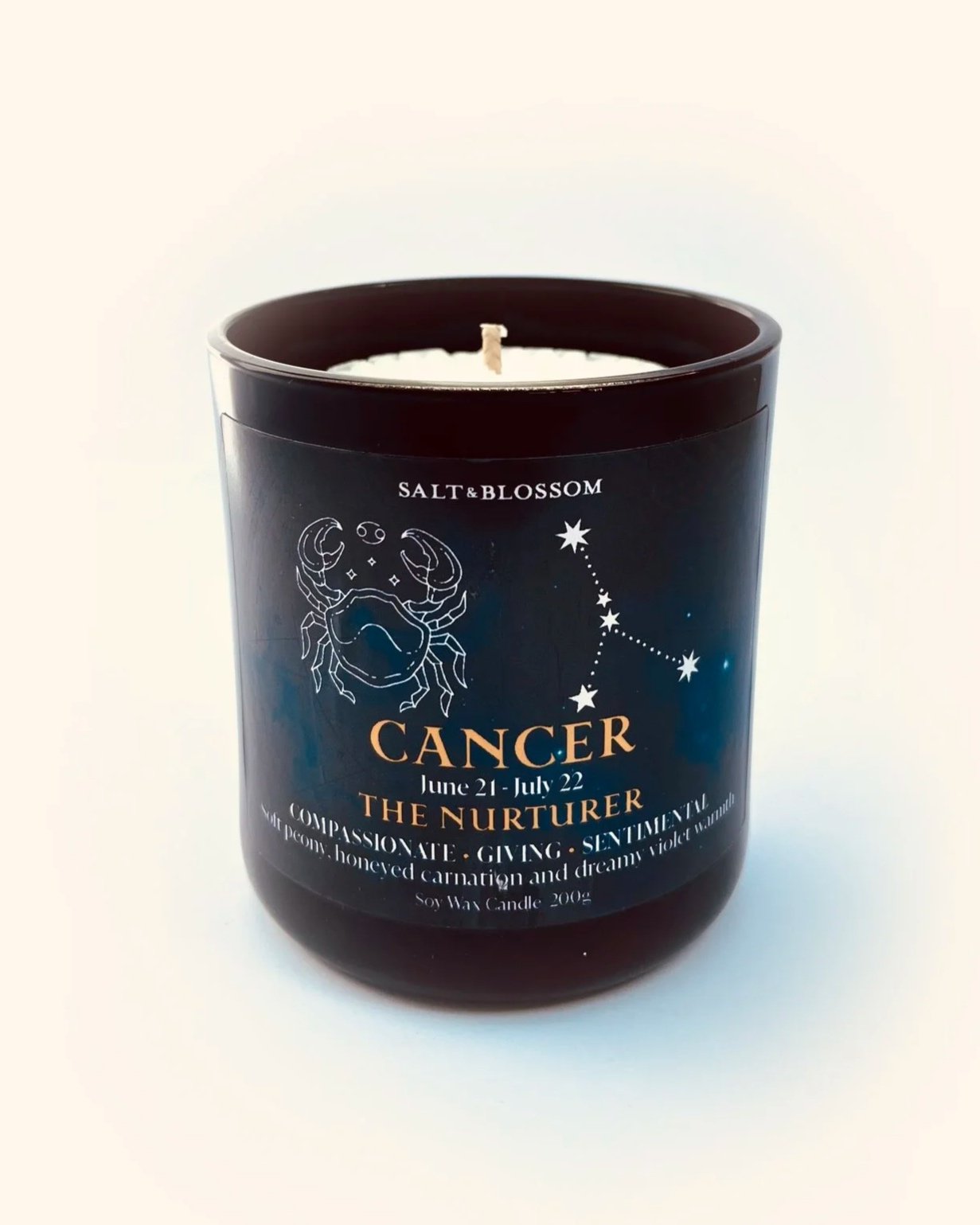 A black scented candle labeled 'Cancer The Nurturer' with a zodiac crab symbol, date range June 21 - July 22, decorated with star constellations and text about compassion, giving, and sentimentality.
