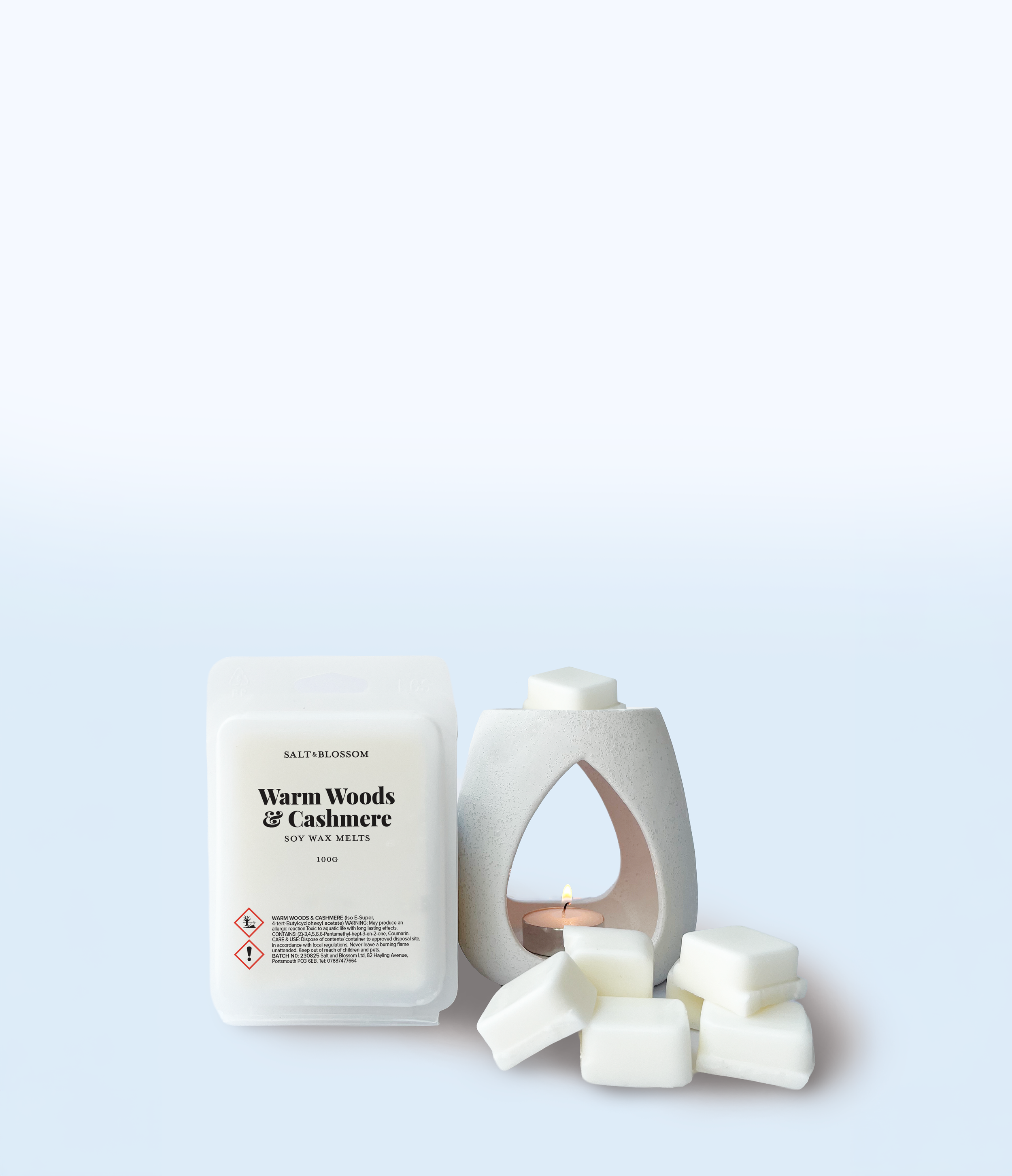 A bottle of soy wax melts labeled warm woods and cashmere, surrounded by white wax cubes and a modern decorative candle holder with a lit candle inside.