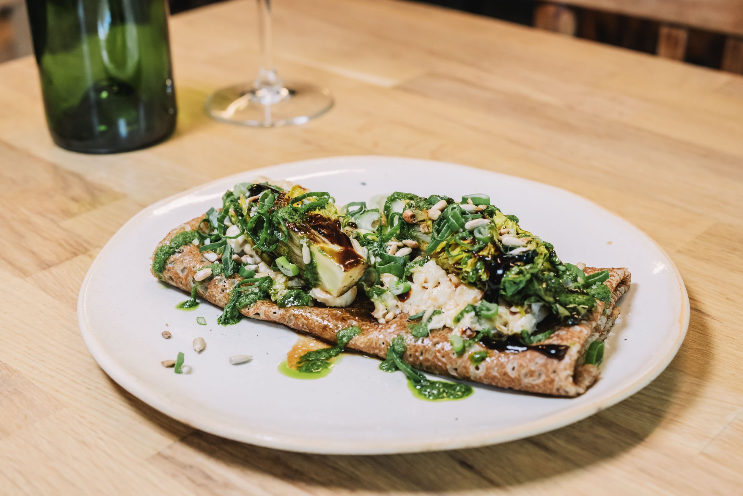 Assiette de crêpe salée garnie de légumes verts, fromage et sauce, sur une table en bois avec un verre et une bouteille. Image de Yves Daniel.