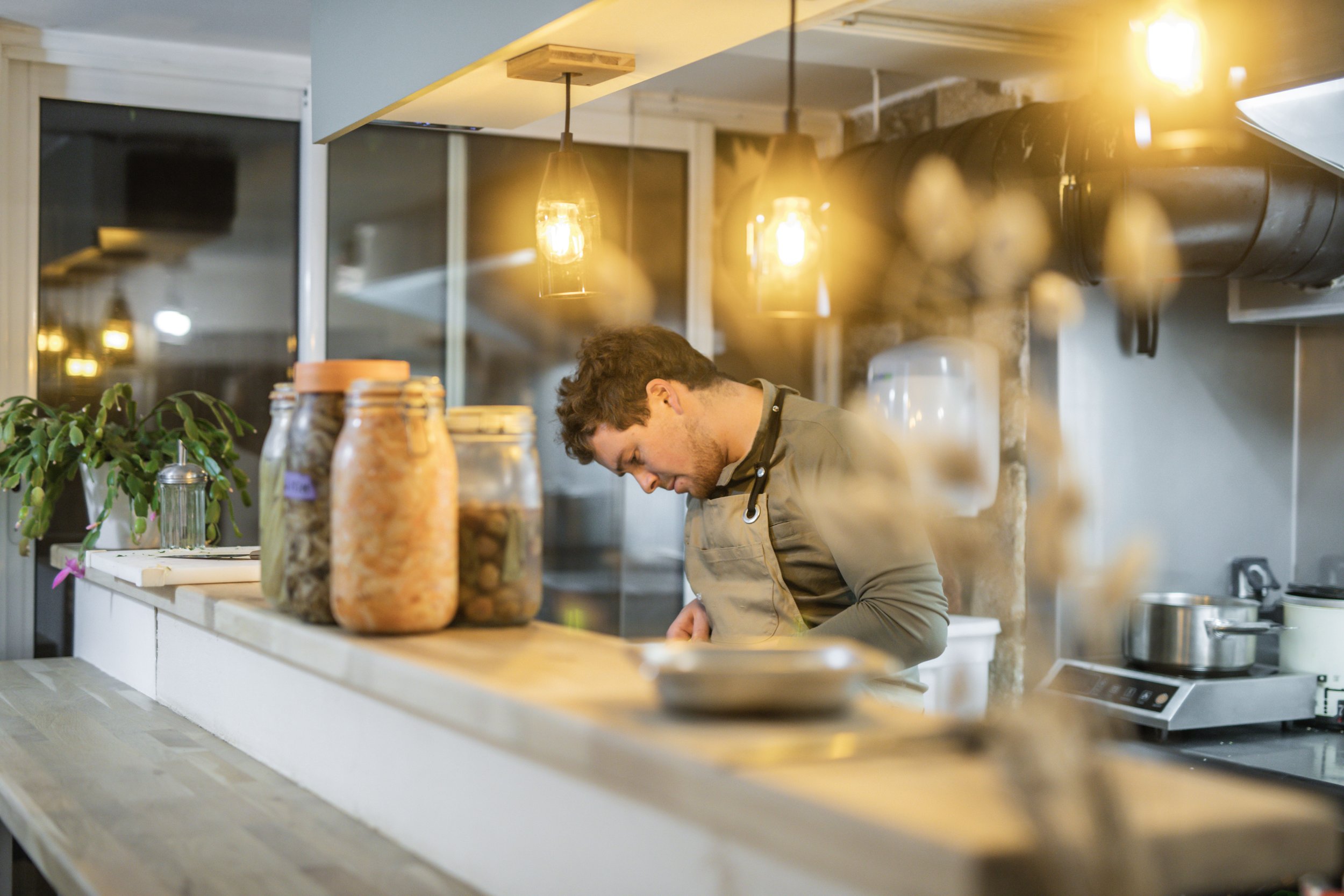 Homme cuisinant dans une cuisine moderne, éclairée par des lampes suspendues avec des bocaux de conserves sur le comptoir. Image de Yves Daniel.