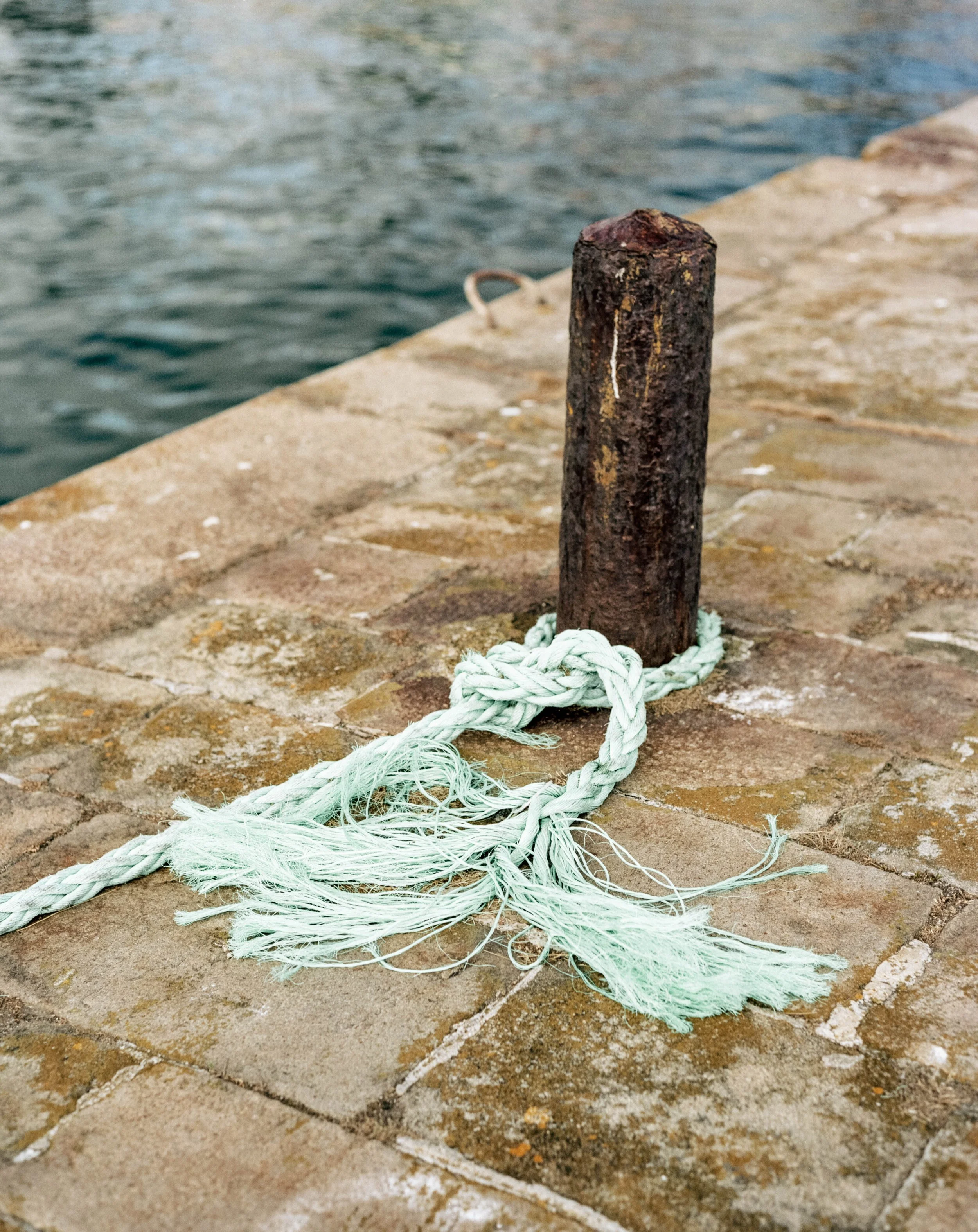 Bollard en bois avec corde attachée sur un quai en pierre près de l'eau. Photo artistique en argentique par Yves Daniel.