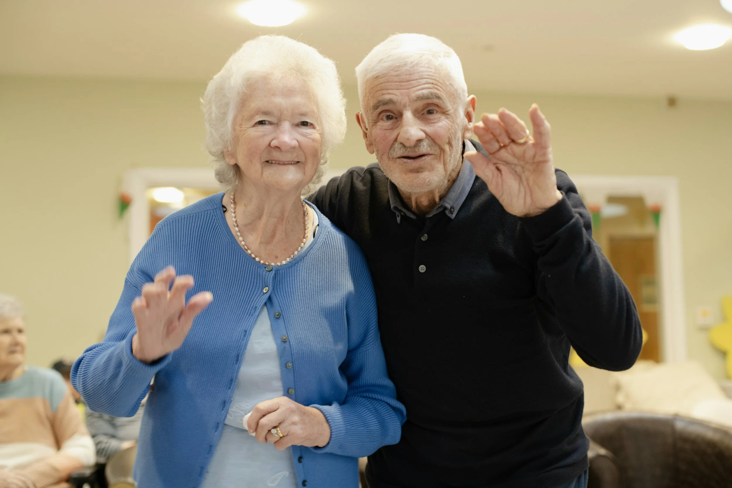 An elderly woman and an elderly man are smiling and waving at a social gathering with other people sitting in the background.