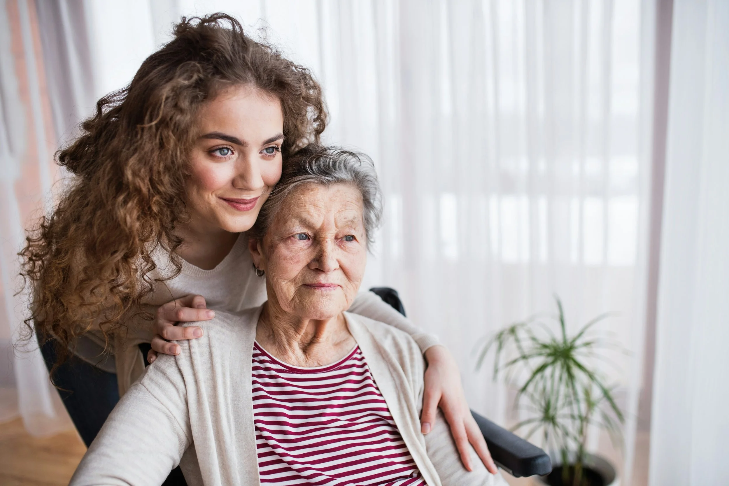 A young woman with curly hair and a light complexion, smiling and leaning over an older woman with gray hair and a light complexion, who is sitting in a wheelchair. The setting appears to be indoors with sheer white curtains and a plant in the background.