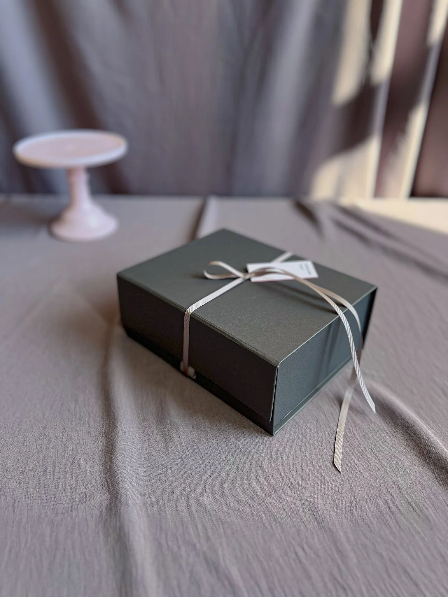 Luxury Grey Gift Box tied with a grey ribbon and Scholars Cakes tag, for the Valentines Day, Mother’s Day, or Birthday Gift Box Bundtette gifting idea - sitting against a grey material studio backdrop with natural light shining in