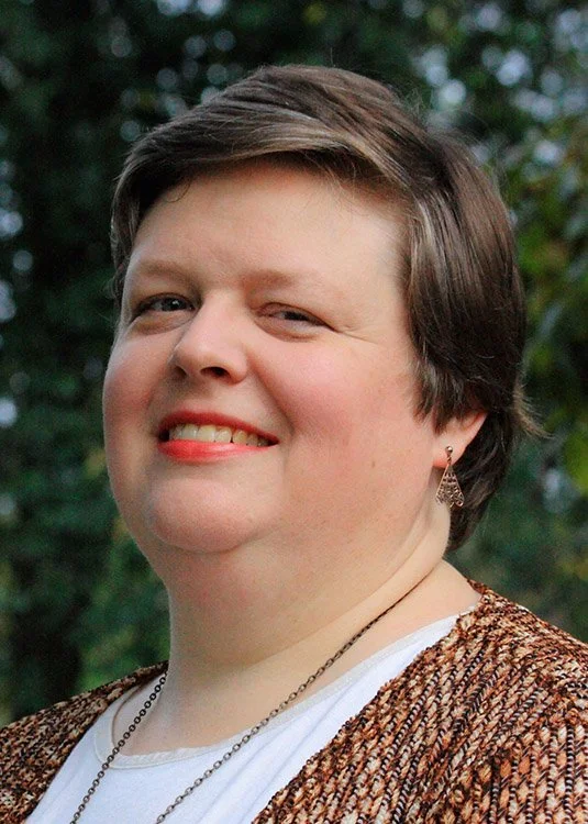 A woman with short brown hair smiling outdoors, wearing earrings, a white shirt, and a brown patterned cardigan, with trees in the background.