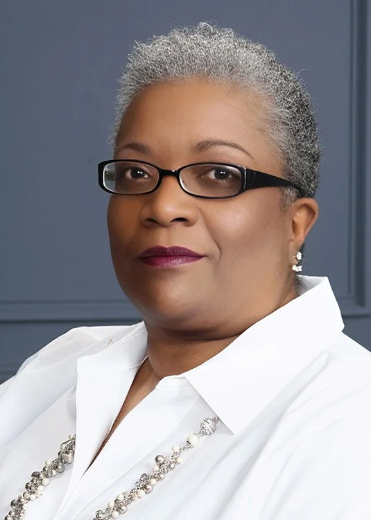 A confident woman with short gray hair, wearing glasses, a white blouse, and a pearl necklace, posing against a blue background.