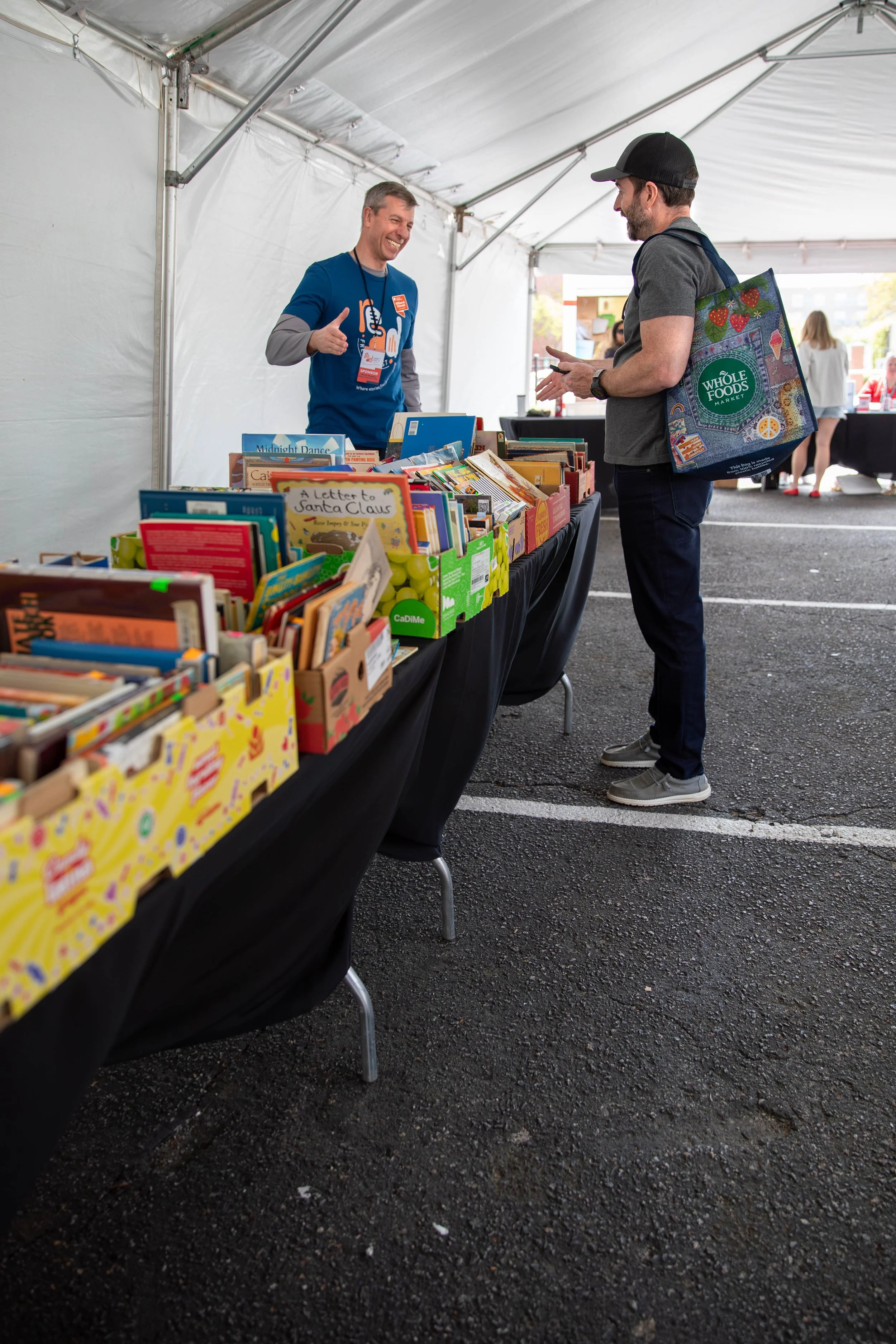 A man shopping at a book sale booth under a tent, smiling and talking to a vendor who is giving a thumbs-up.