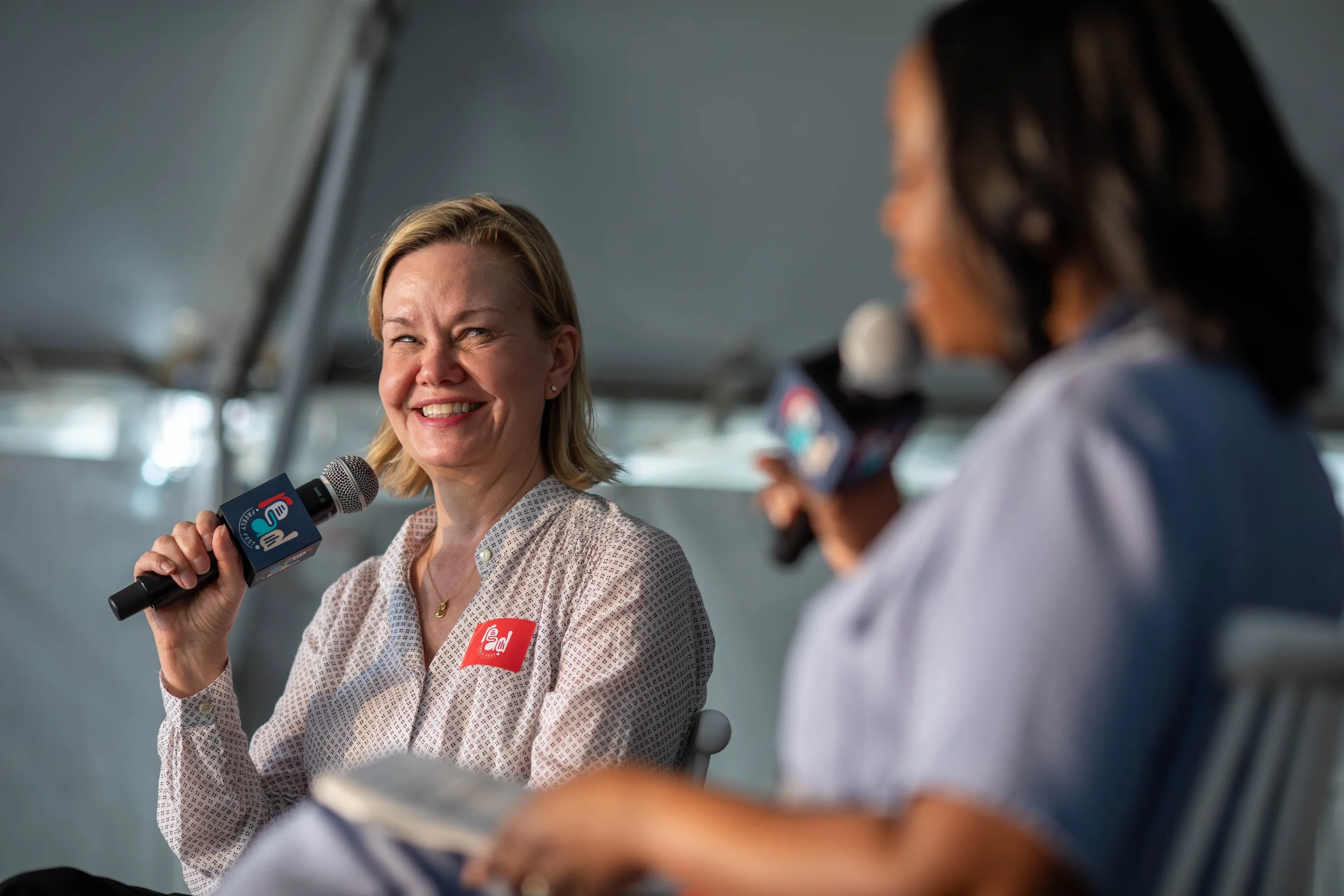 Two women are participating in an interview or panel discussion under a tent. The woman on the left is smiling, holding a microphone, and wearing a white shirt with a red badge that says "End." The woman on the right is partially visible, holding a m