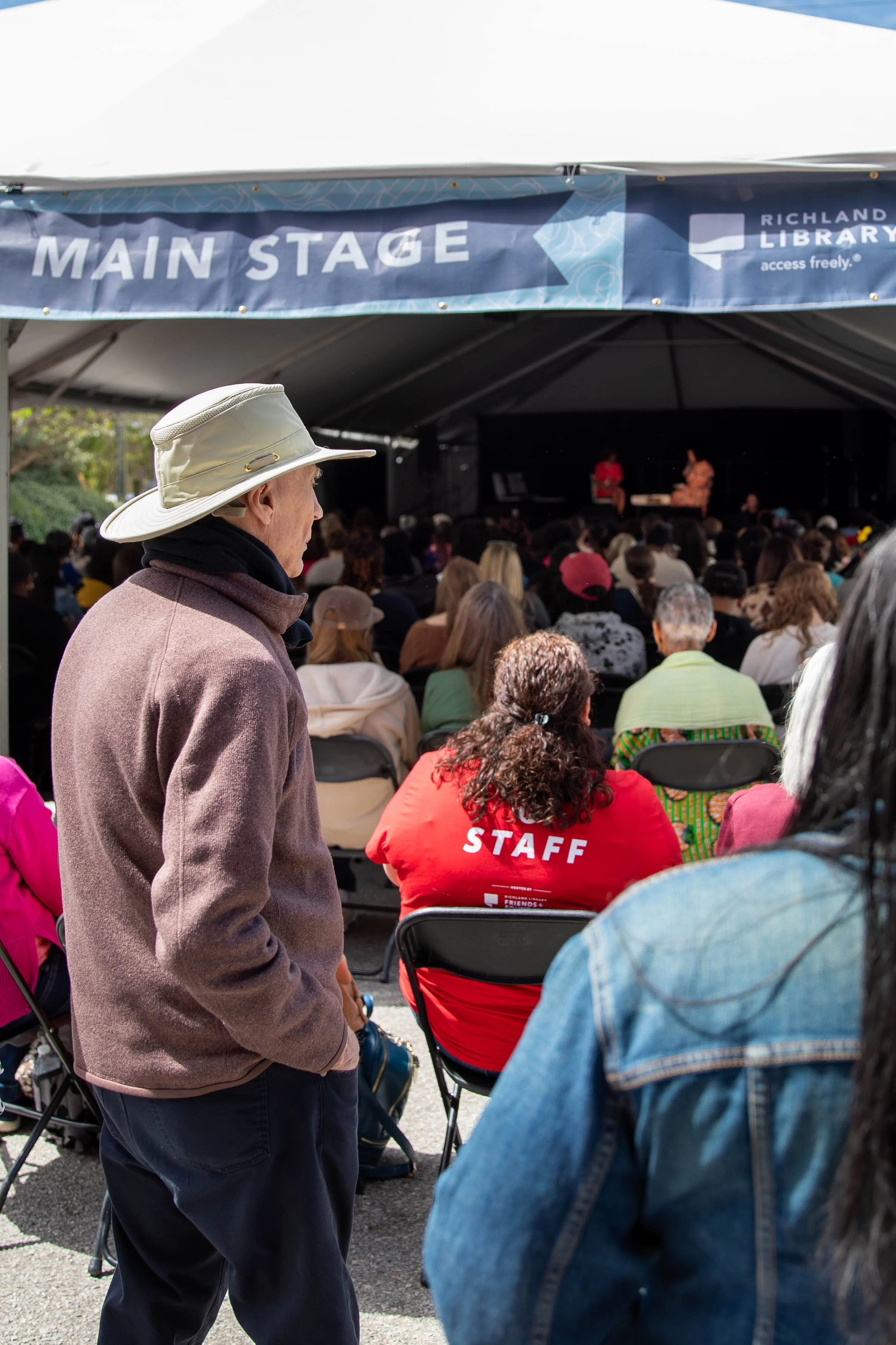 An outdoor event with a standing man wearing a beige wide-brimmed hat and brown jacket, observing a seated audience facing a stage under a blue canopy. The stage has performers, and a sign above reads 'MAIN STAGE,' near a sign for 'Richland Library'.