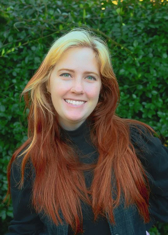 A young woman with long, wavy red hair and fair skin smiling outdoors against a background of green foliage.