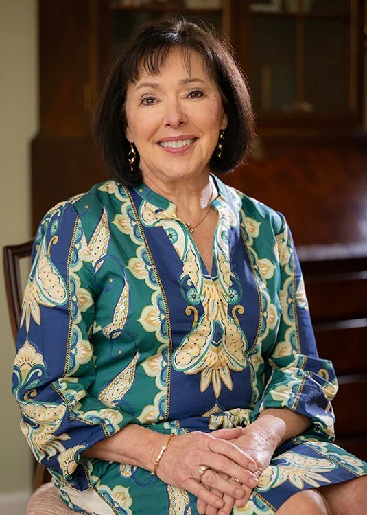 A smiling woman with dark hair sitting in a room with wooden furniture, wearing a colorful patterned dress and jewelry.