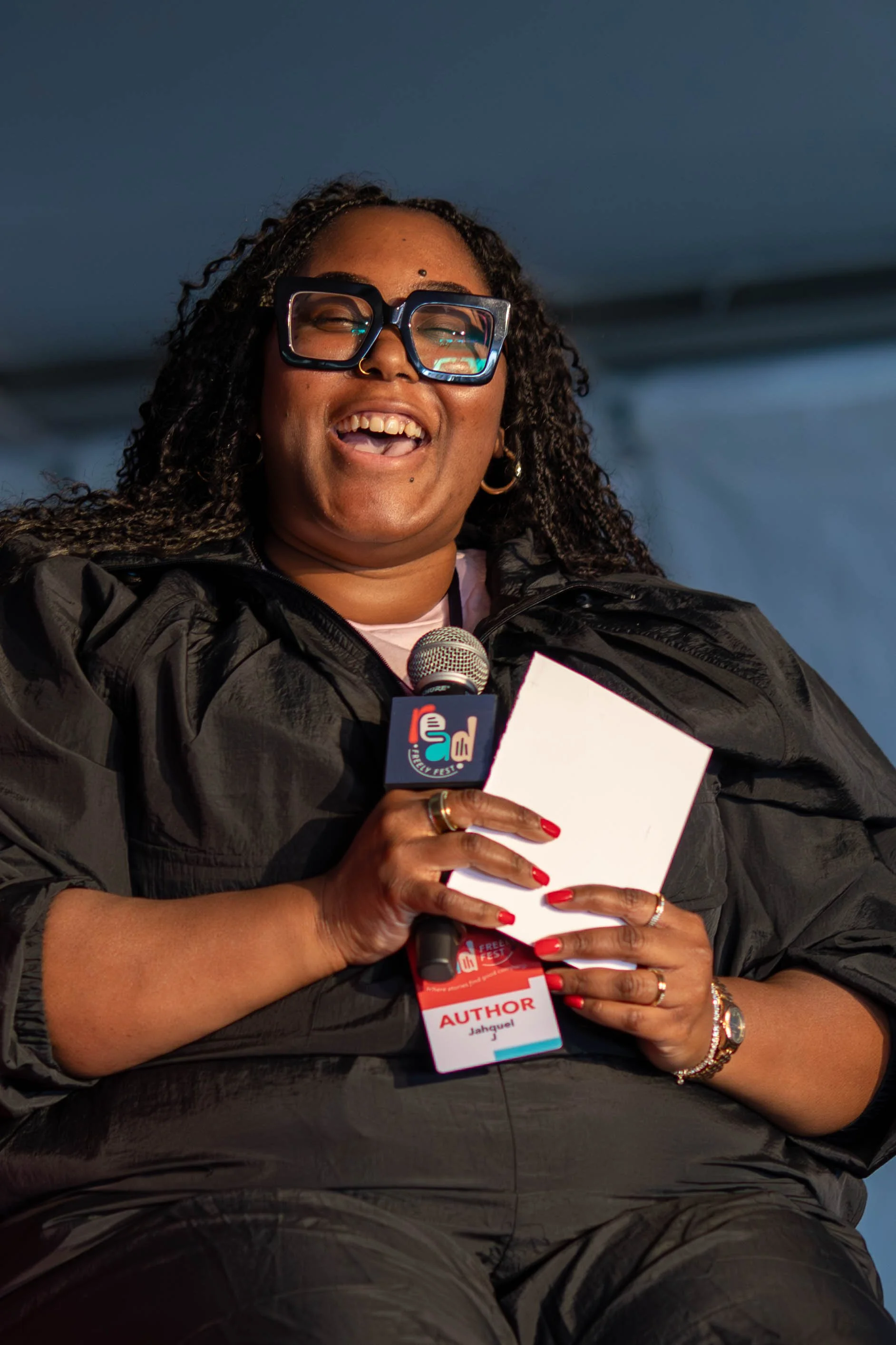 A woman with curly hair, glasses, and gold jewelry smiling and holding a microphone and a notepad, wearing a black jacket and an author badge, speaking at an event.