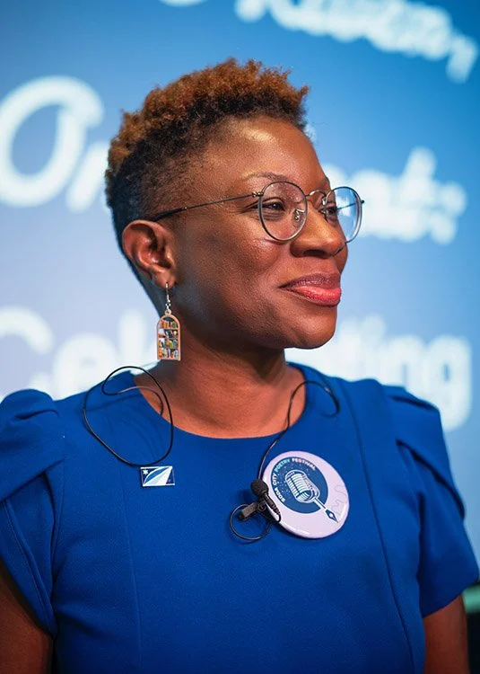 A woman with short curly hair, glasses, and earrings, wearing a blue dress or uniform, standing in front of a blue background with white writing.