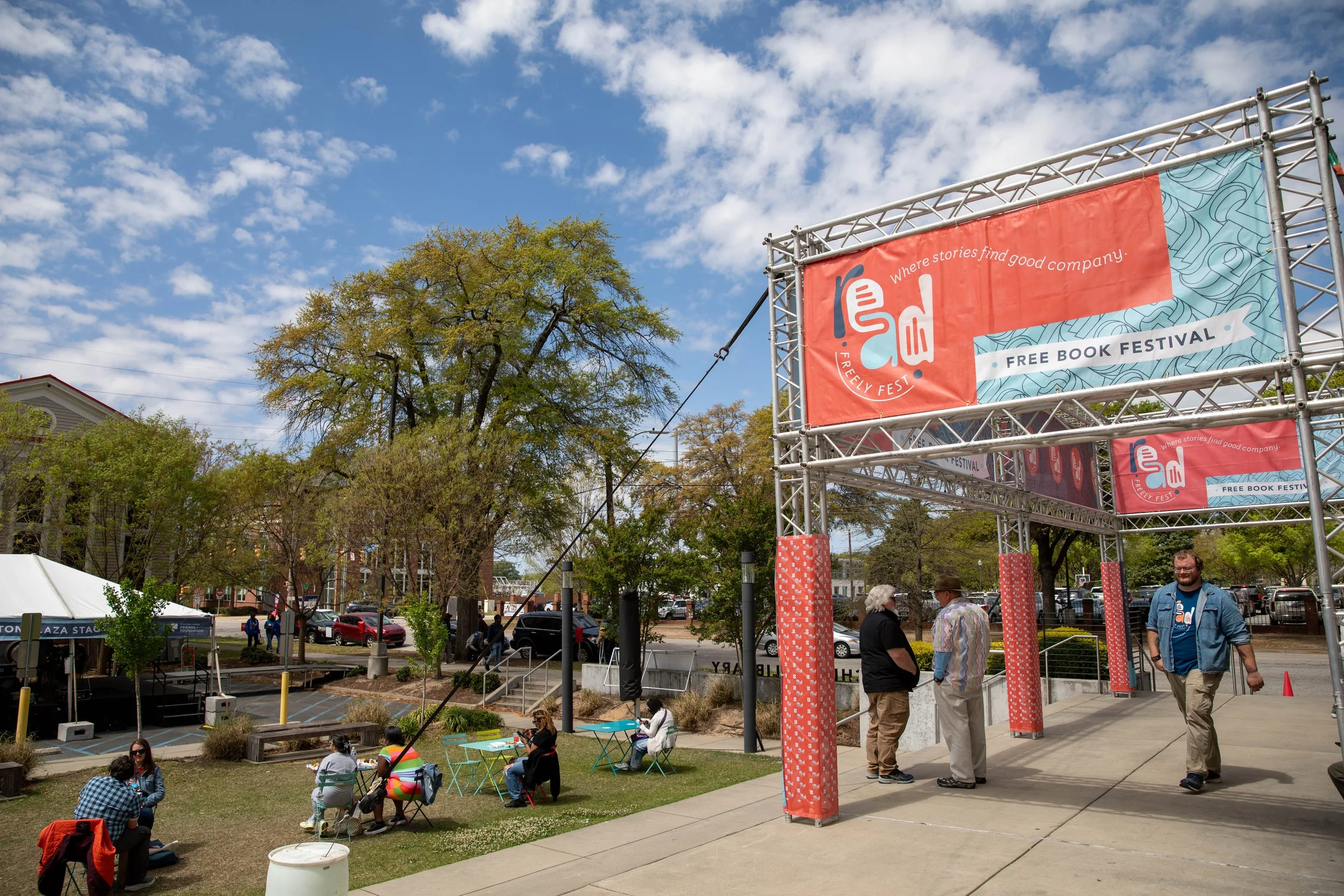People enjoying a sunny day at a free book festival under a large red and blue sign that reads 'Where stories find good company,' with a smaller banner saying 'Free Book Festival'.