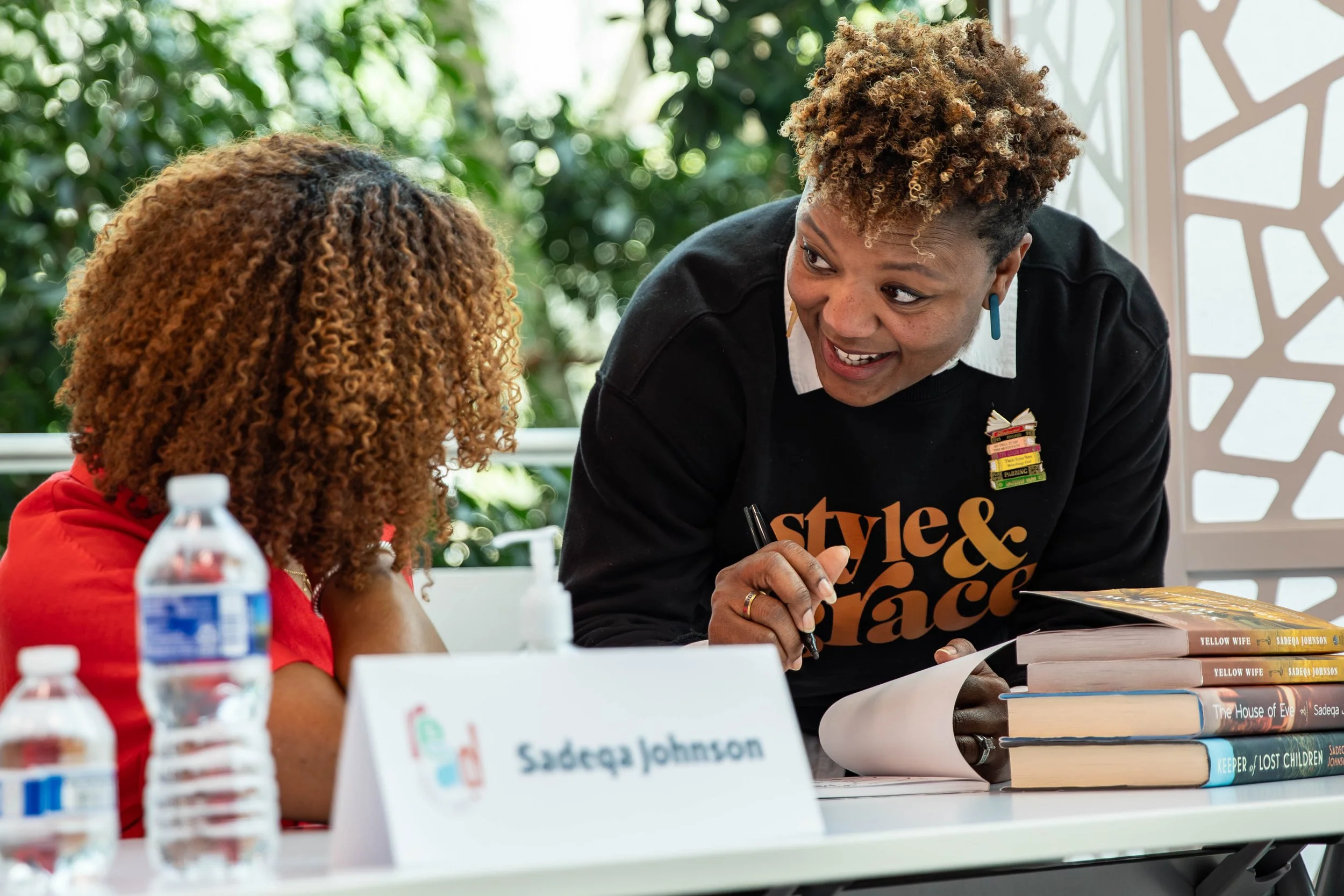 Woman engaging with a young girl at a signing event, sitting at a table with books, water bottles, and a nameplate reading 'Sadeqa Johnson'. The woman is smiling, holding a pen, and wearing a black sweatshirt that says 'styles & grace' with books sta
