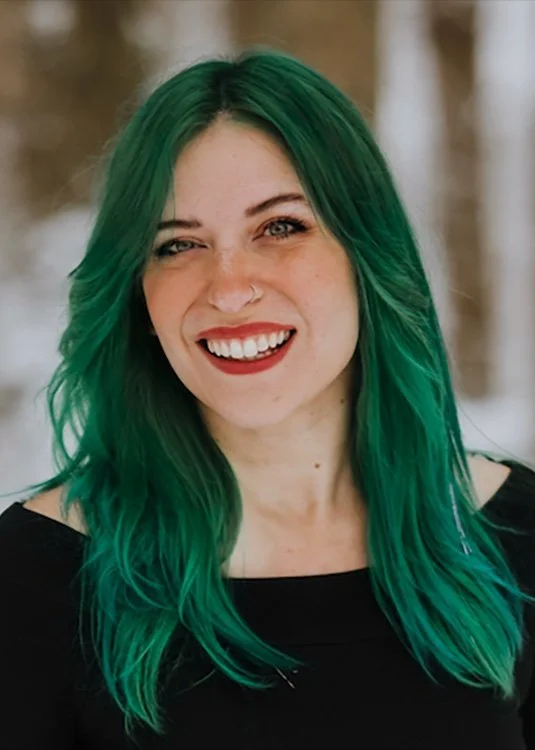 A woman with long green hair, smiling with red lipstick, wearing a black top, outdoors with trees in the background.