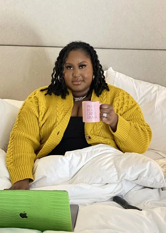 A woman with dark curly hair wearing a yellow cardigan and black top, sitting in bed and holding a pink mug that says 'Pray Read Grind Drank'.