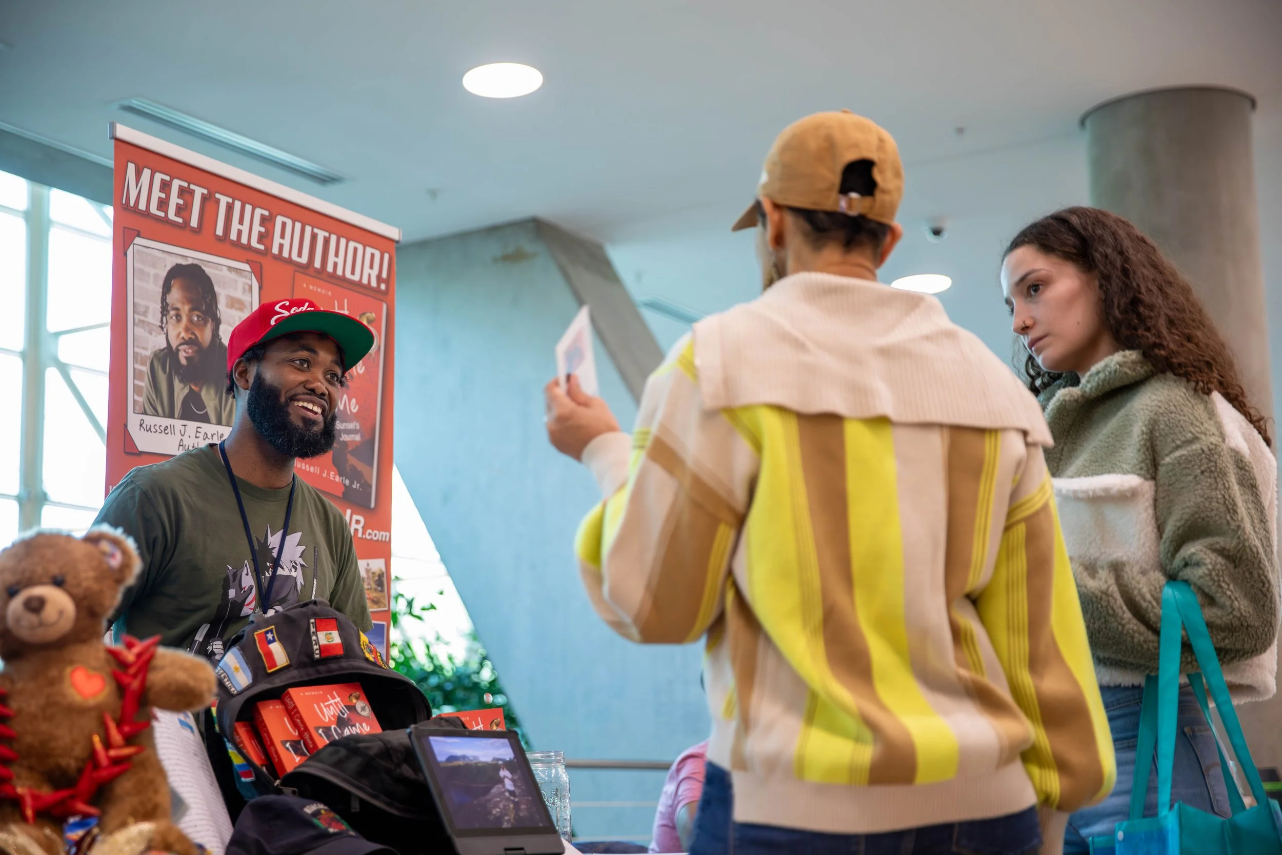 A man with a beard wearing a red cap and green shirt stands at a booth with a sign that reads 'Meet the Author!' and a photo of Russell J. Earle Jr. The booth has stuffed bears and books on display. Two women are talking to him, one in a yellow strip