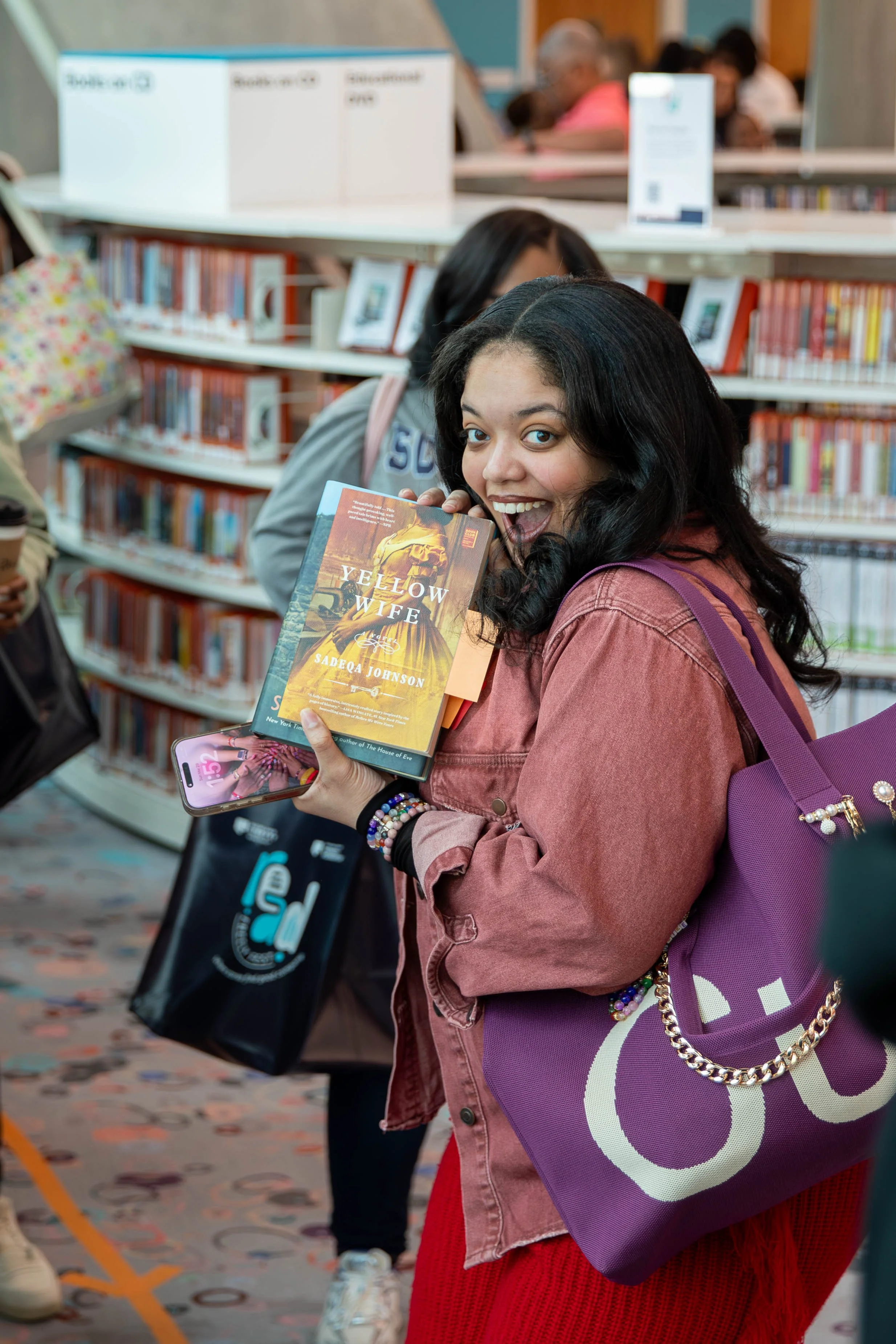 A young woman in a denim jacket and red skirt holding a paperback book titled 'Yellow Wife' by Sadeqa Johnson at a library, smiling excitedly at the camera, with other people browsing books behind her.