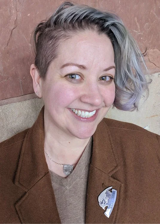 A woman with short, styled hair that is dark on one side and light purple on the other, smiling and wearing a brown jacket with a pin and a necklace, against a background of a pinkish wall and beige surface.