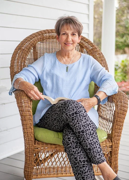 An older woman with short hair, wearing a light blue shirt and leopard-print pants, sitting on a wicker chair with a green cushion on a porch, holding an open book and smiling at the camera.