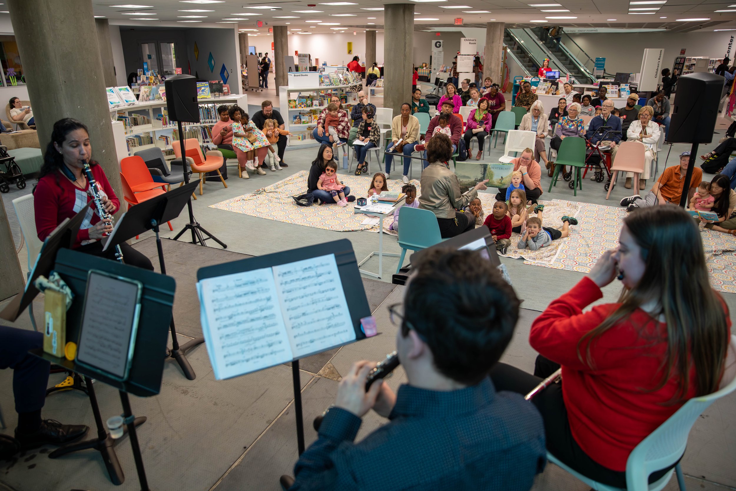 Children and adults attending a library storytelling event with musicians playing wind instruments and a storyteller reading to kids sitting on blankets.