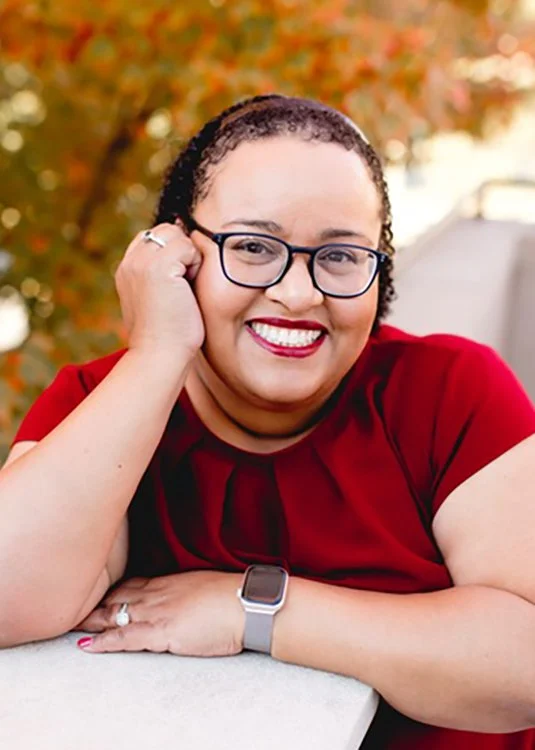 A smiling woman with glasses and short curly hair, wearing a red top, resting her head on her hand outdoors with autumn-colored trees in the background.