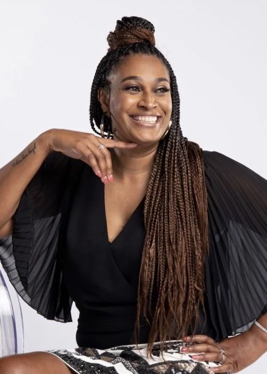 A woman with long braided hair wearing a black pleated dress, smiling and posing with one hand near her face and the other on her hip.