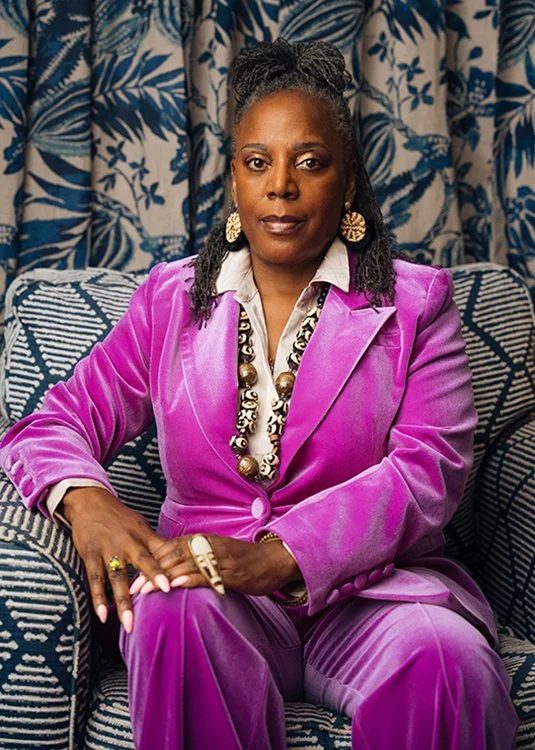 A woman sitting on a sofa with a patterned background, wearing a purple suit, large earrings, and a chunky necklace.