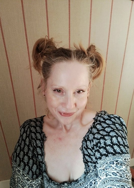 A woman with light skin and blonde hair styled in two messy buns, smiling at the camera, wearing a black and white patterned top, standing in front of a beige and red striped wall.
