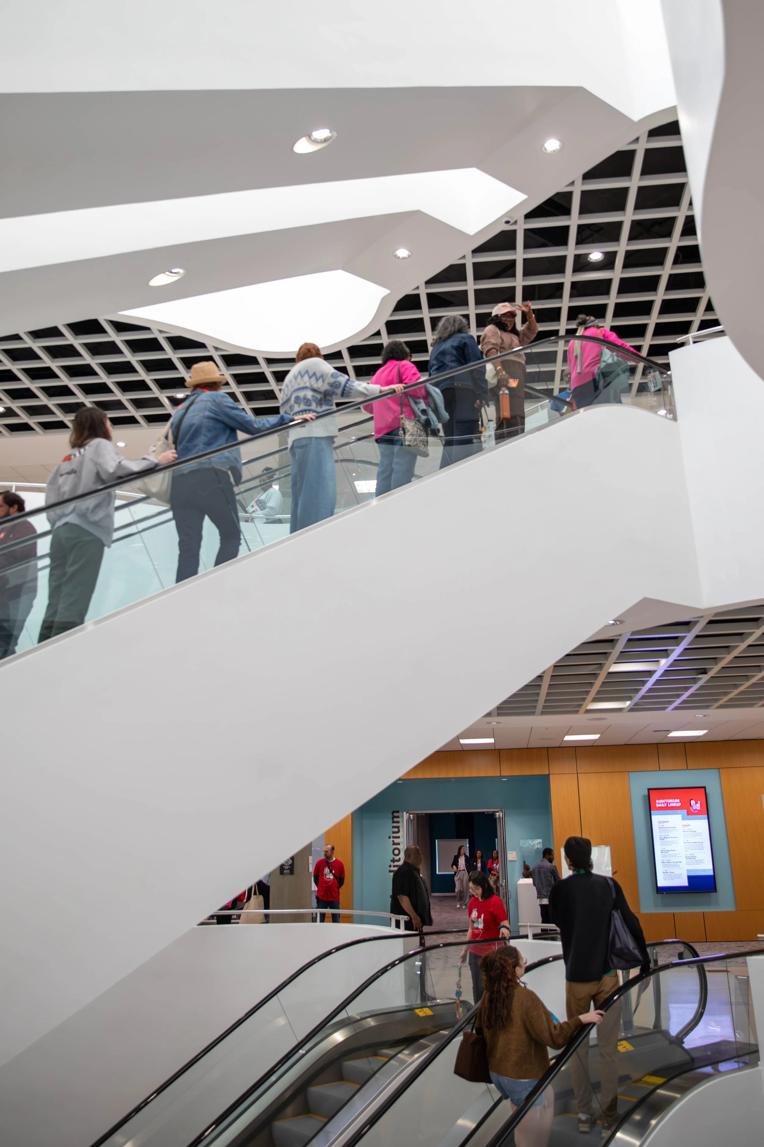 People walking on escalators inside a modern airport terminal with a white ceiling featuring geometric cutouts and a digital display board.