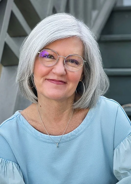 A smiling elderly woman with short gray hair and glasses, wearing a light blue top and a necklace, posing outdoors near stairs.