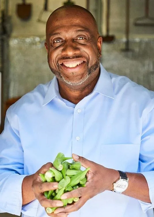 A smiling man holding a bunch of celery in a kitchen with a blurred background.