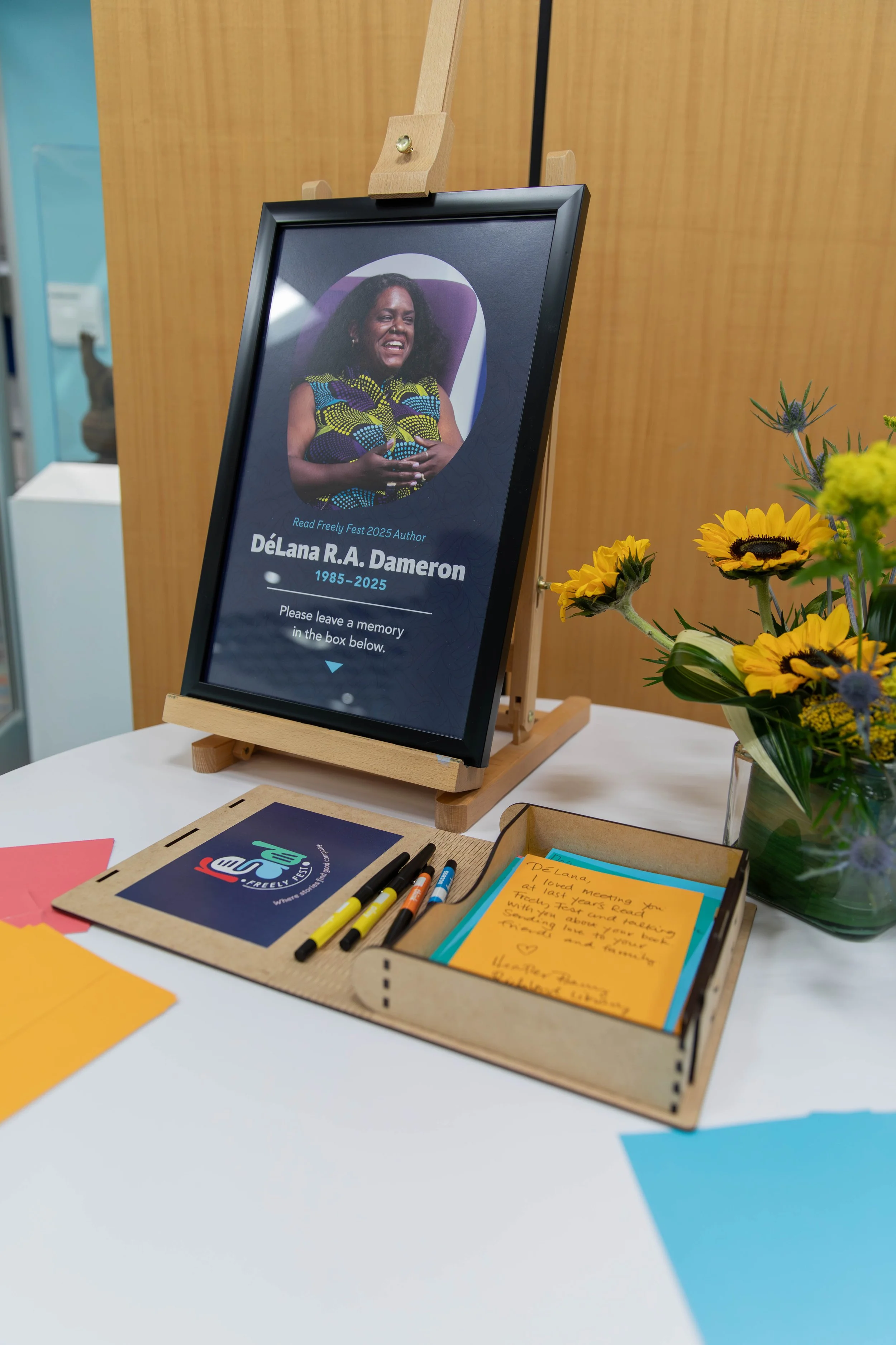 A memorial display for DéLana R. A. Dameron, including a framed digital tribute, yellow flowers, and handwritten notes on a desk.