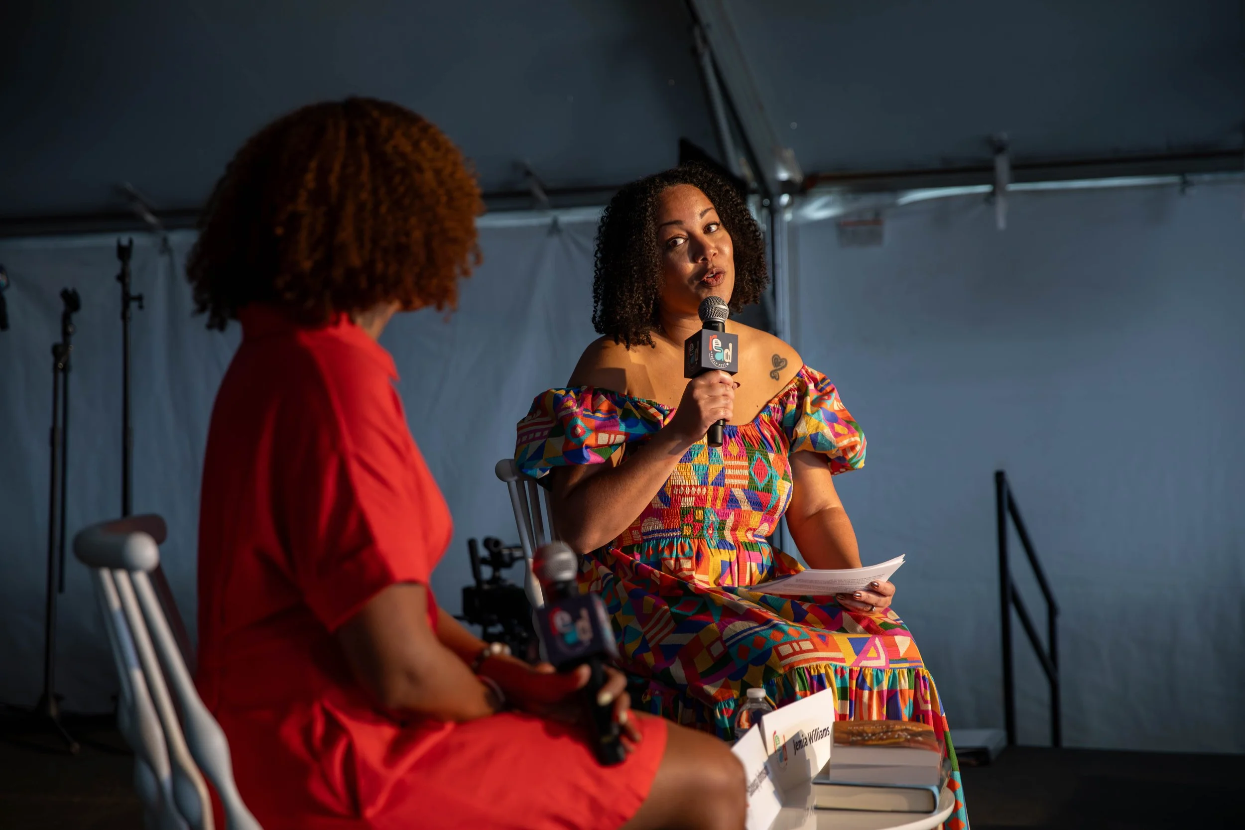 Two women engaging in a discussion on stage at a panel event inside a tent, with microphones and books. One woman is wearing a colorful off-shoulder dress, and the other is dressed in a red outfit.
