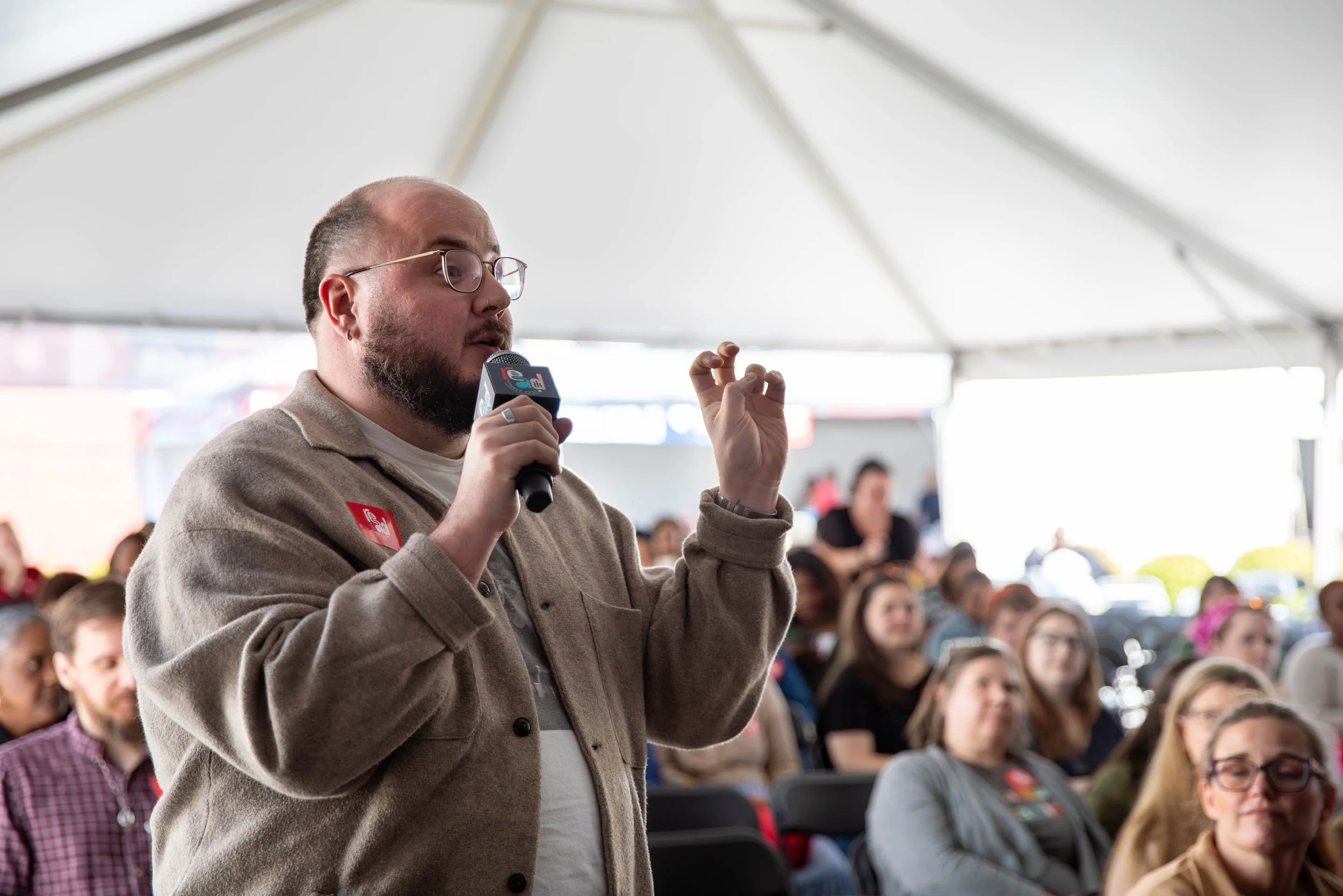 A man with glasses and a beard speaking into a microphone at an outdoor event under a large white tent, with a seated audience of diverse people listening.