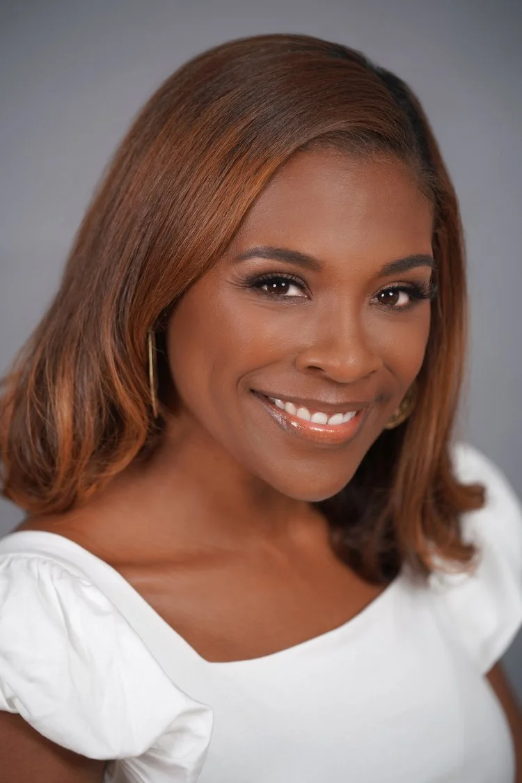 A woman with straight brown hair smiles warmly, wearing a white top and gold hoop earrings against a gray background.