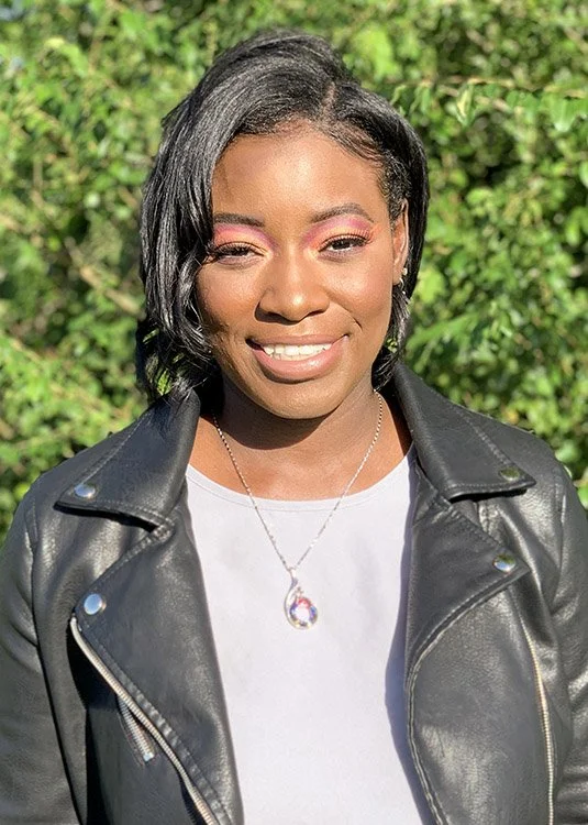 A woman with short black hair, wearing pink eyeshadow, a white shirt, a black leather jacket, and a necklace with a pendant, standing outdoors with green foliage in the background. She is smiling and winking.