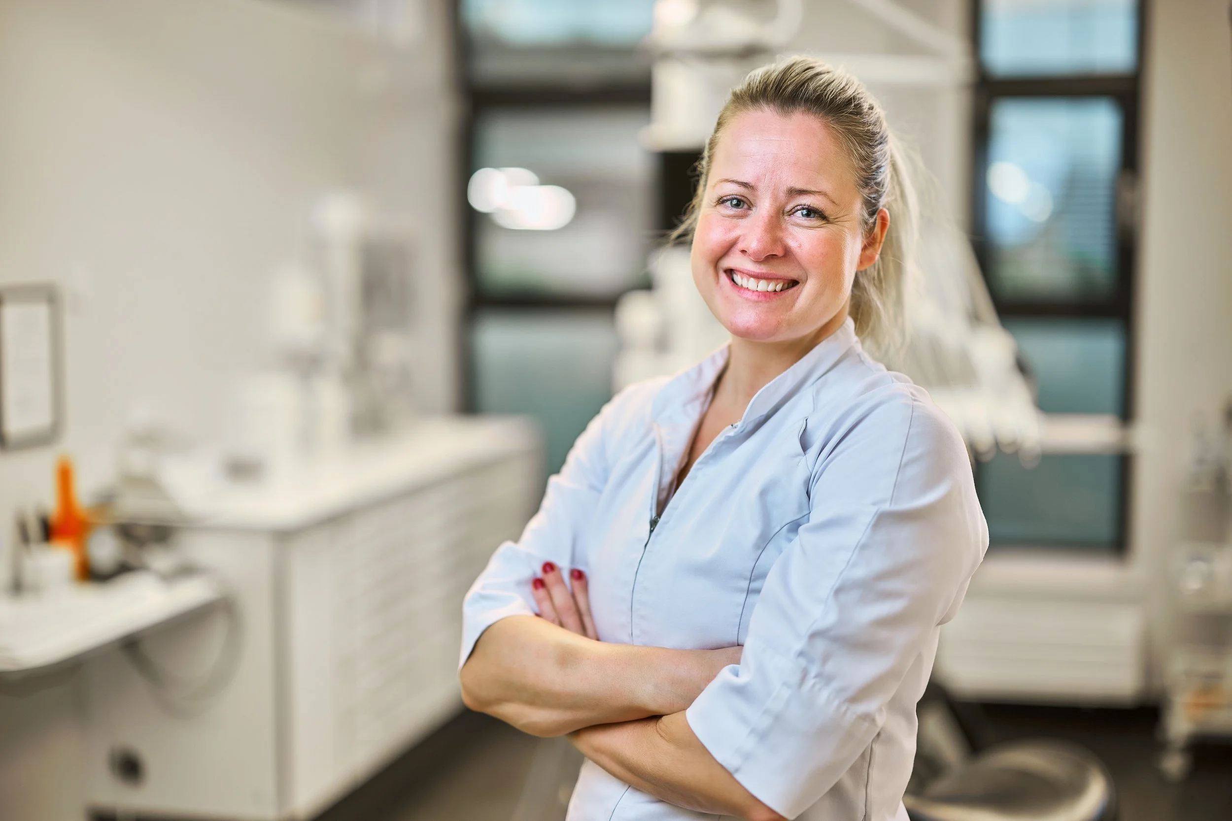 En smilende kvindelig tandlæge i en tandlægeklinik, iført hvidt klinisk tøj med armene over kors, foran tandlægeudstyr.