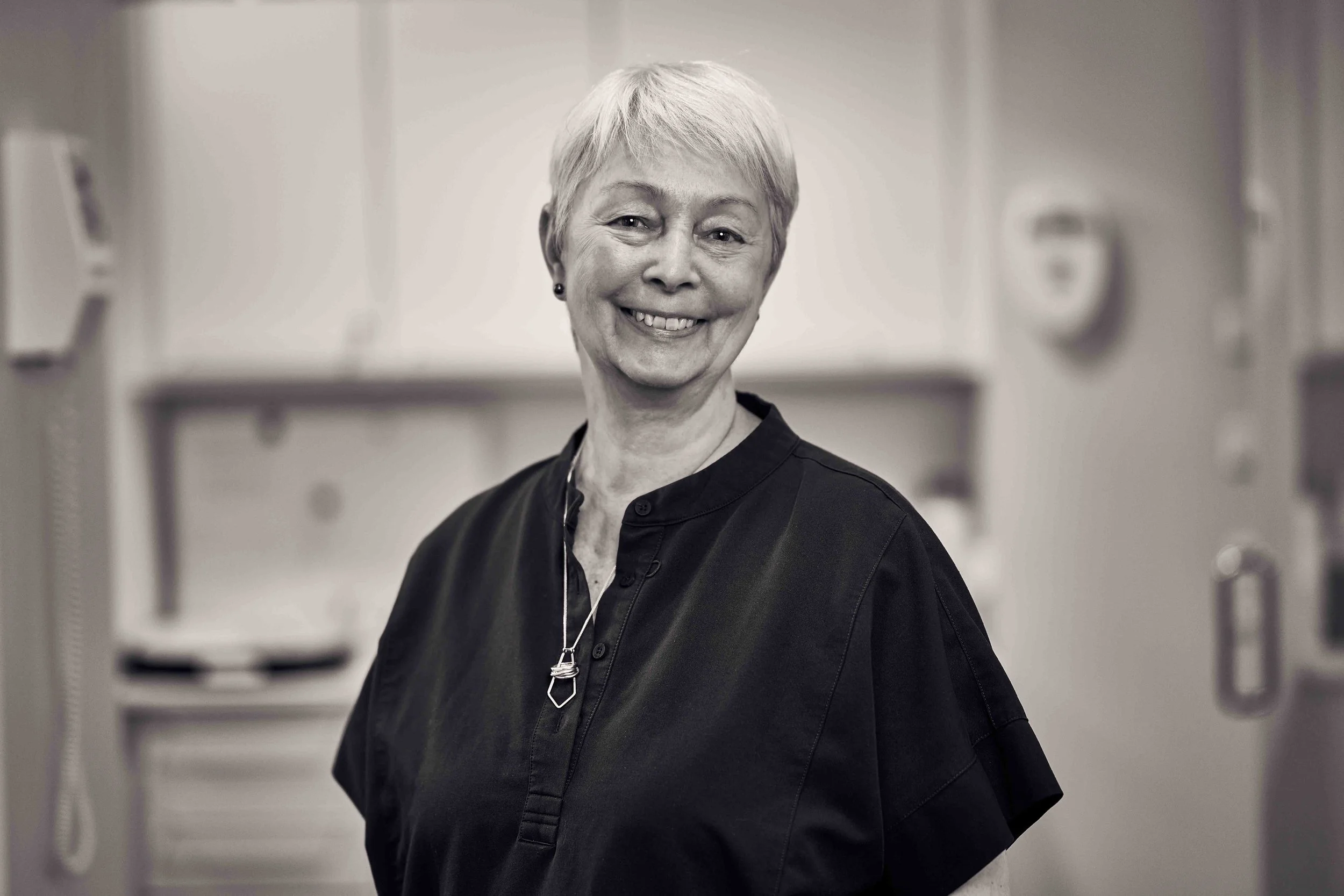 En ældre kvindelig sygeplejerske iført sort uniform, står med hænder bag ryggen, smilende mod kameraet.