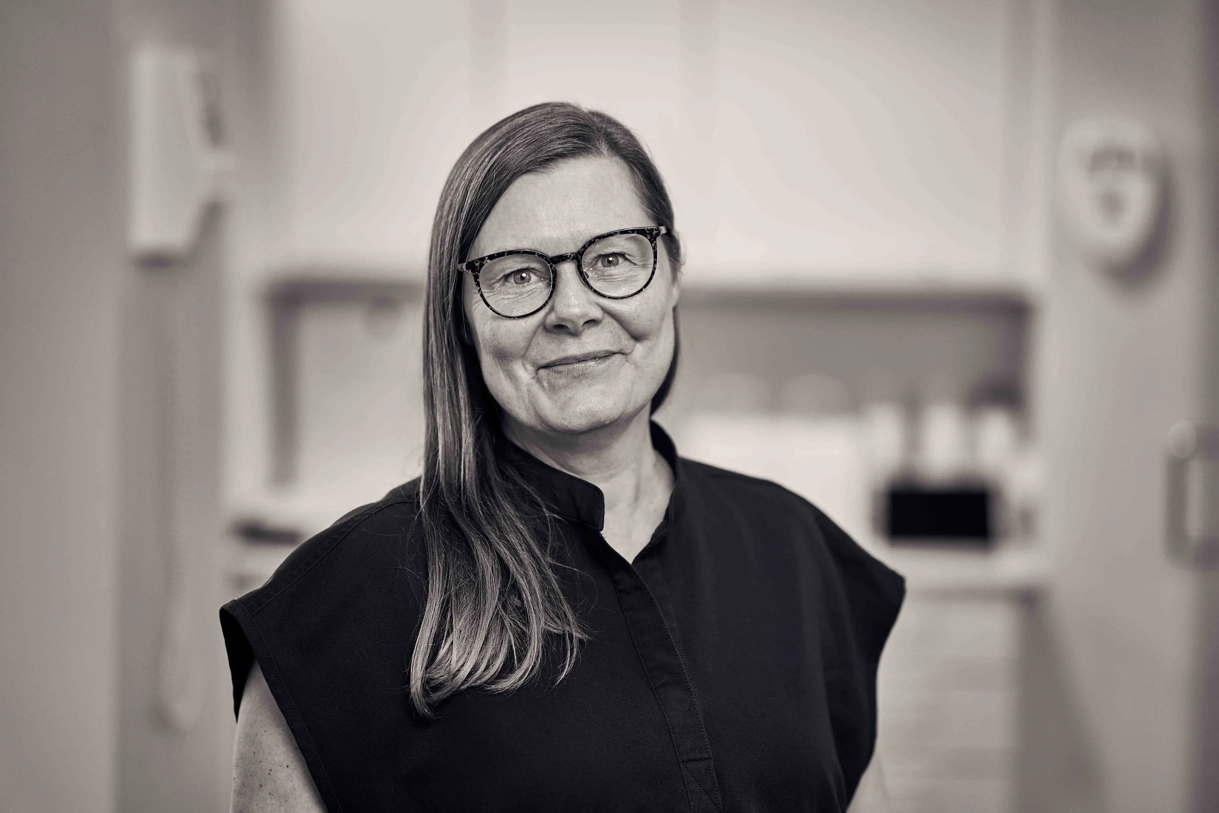 En smilende kvinde med briller, iført en sort jakke og en smal halskæde.