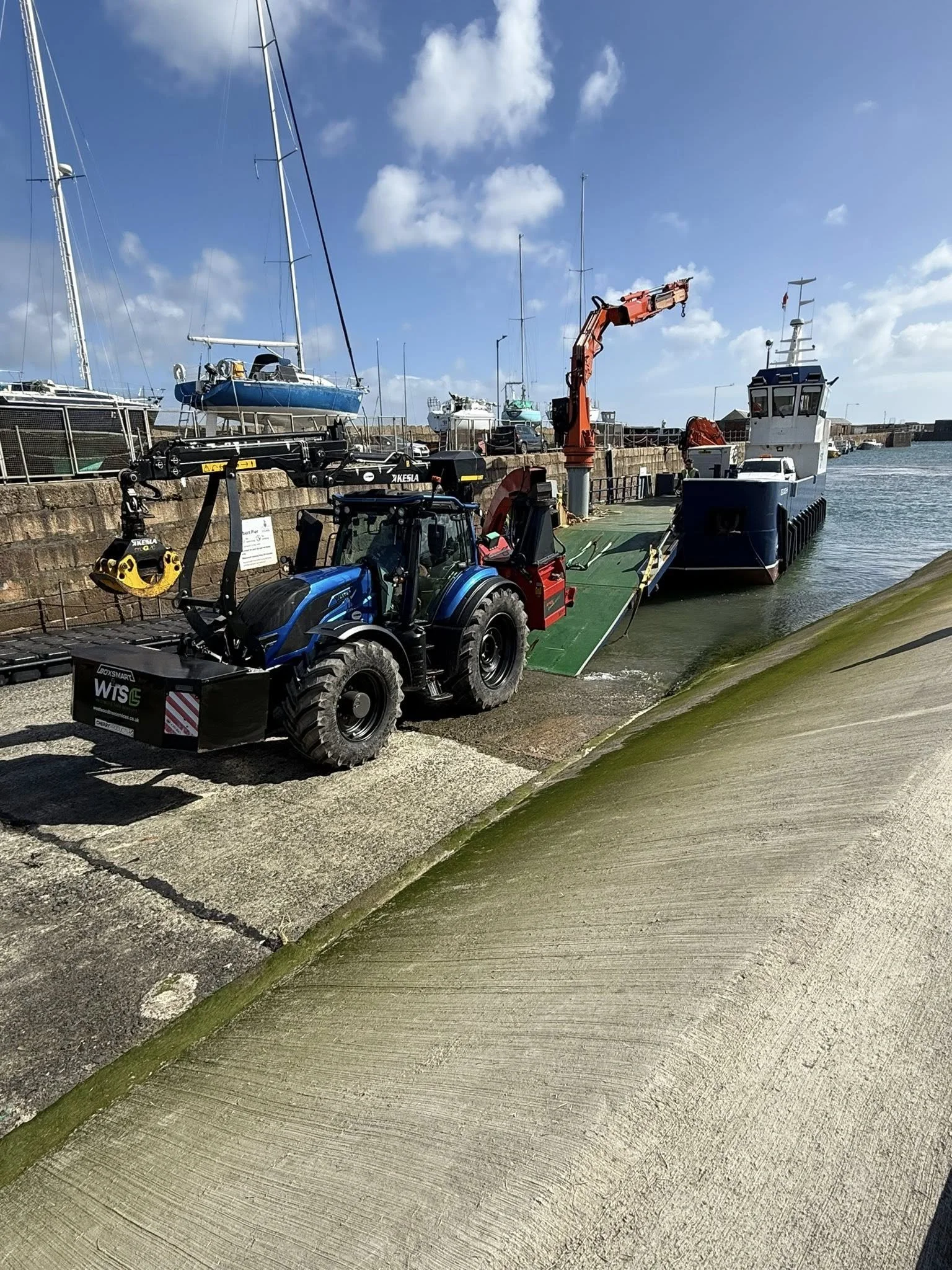 A boat being lowered into the water from a dock with a tractor and a crane. Sailboats and a blue sky with scattered clouds in the background.