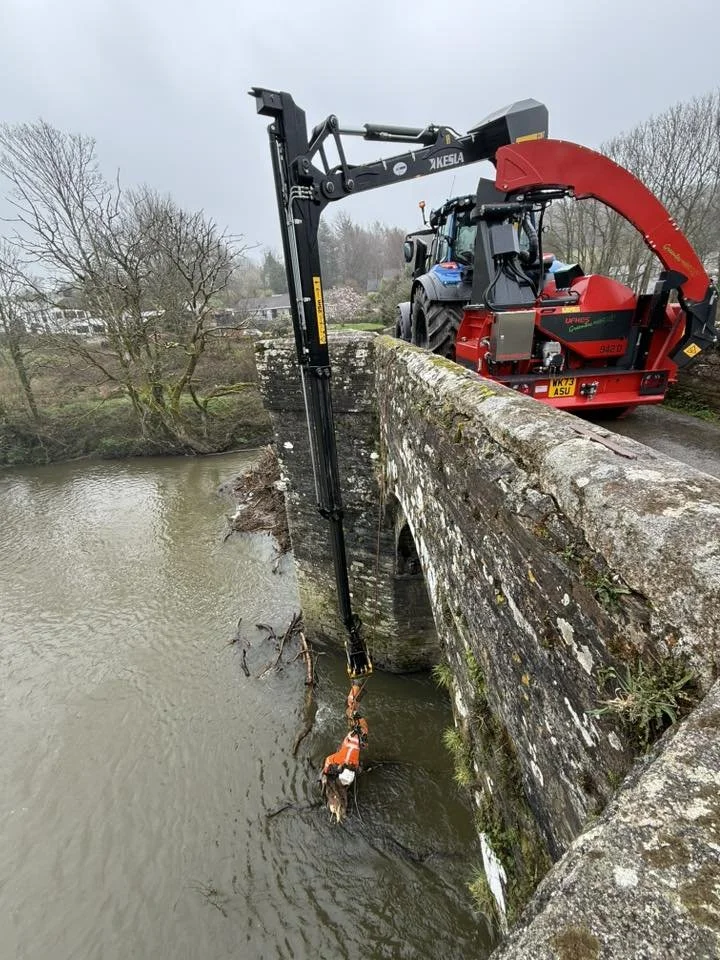 Rescue team using a crane attached to a tractor to save a dog wearing an orange life jacket from a river next to a stone bridge.