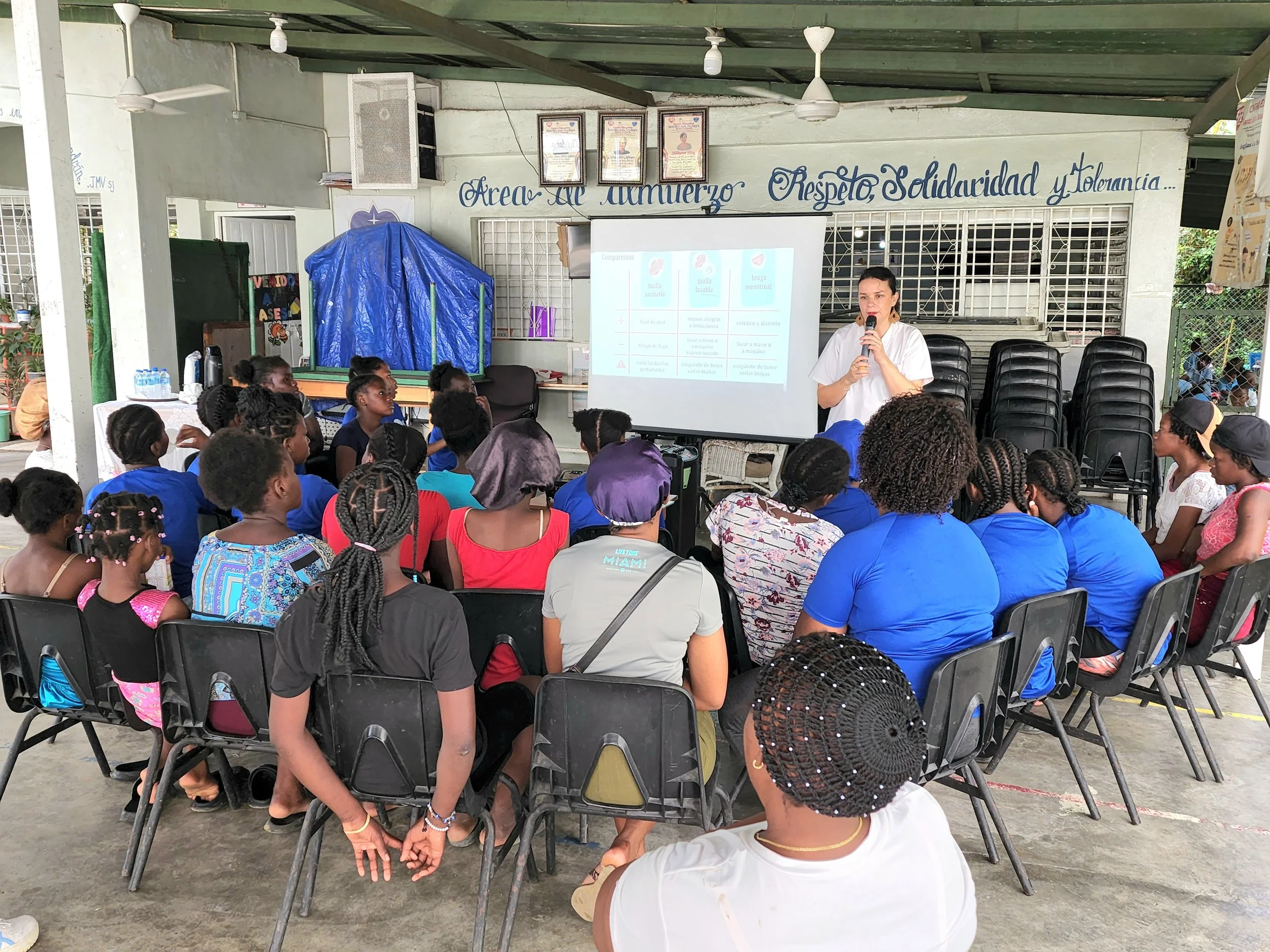 A group of children and adults seated in chairs, attentively listening to a woman giving a presentation in an open-air space with a painted wall and a projector screen.